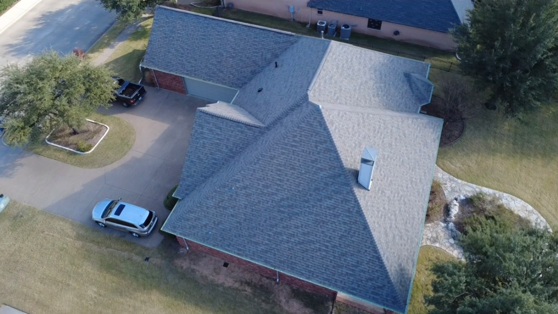 An aerial view of a suburban house with a gray shingle roof, a driveway with parked cars, a backyard with trees and a stone pathway.