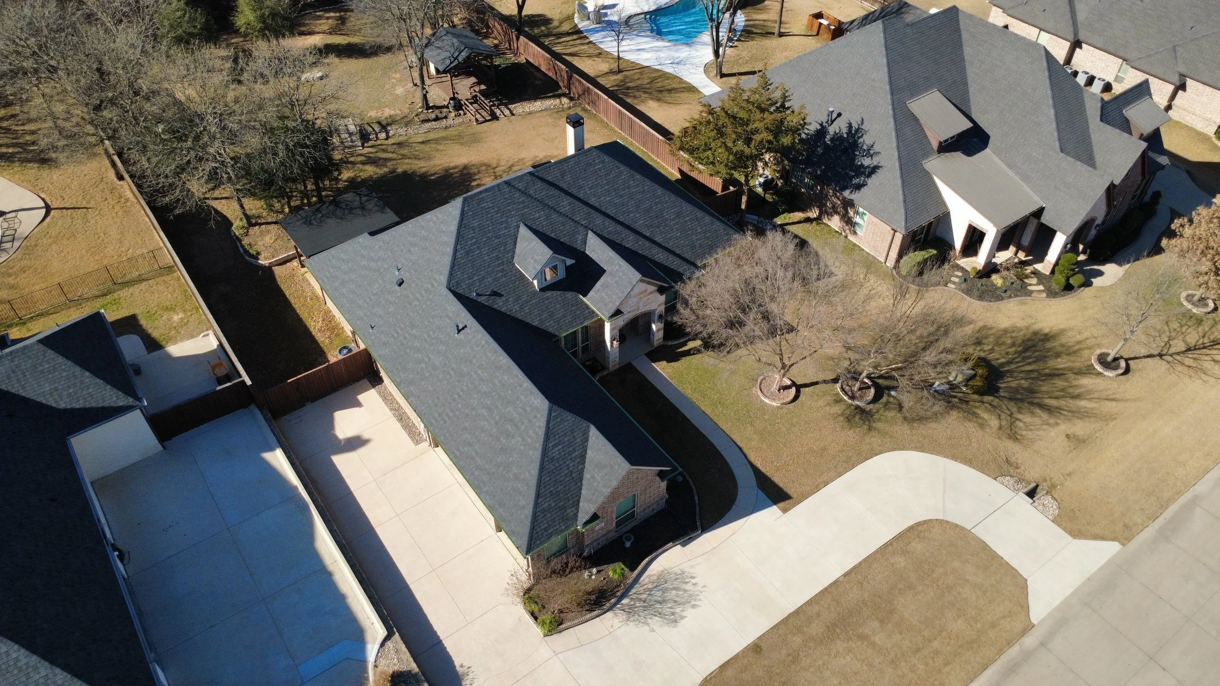 Aerial view of a residential backyard with two houses, a fenced yard, trees, and a swimming pool.