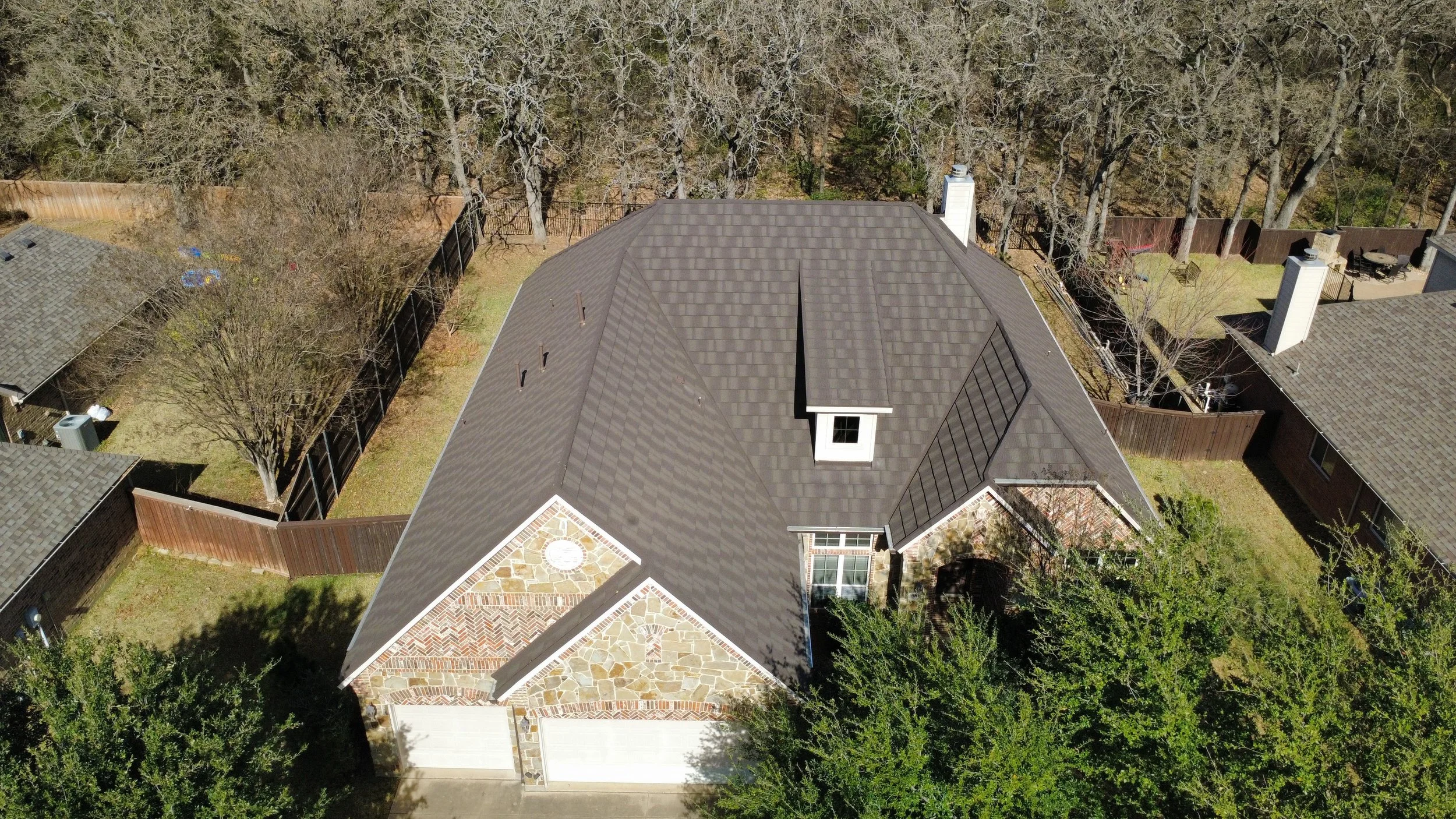 Aerial view of a house with a dark gray shingle roof, a small dormer window, and a stone facade on part of the exterior. The house is surrounded by a fenced backyard with trees and neighboring houses.