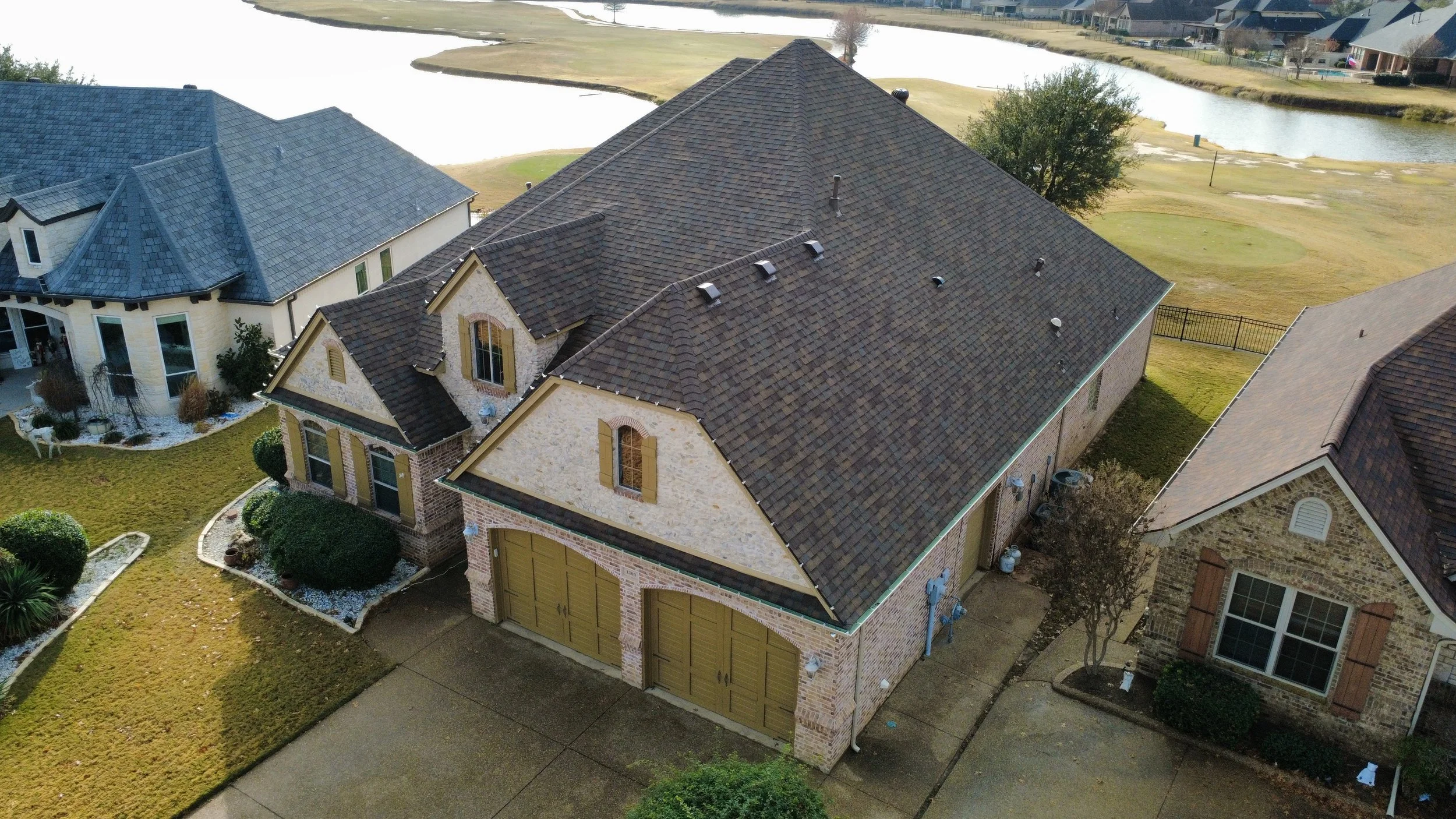 An aerial view of a large suburban house with a brick and stone exterior, a high pitched shingle roof, a two-car garage with yellow doors, and a landscaped front yard. There is a neighboring house with similar architecture and a water body behind the houses.