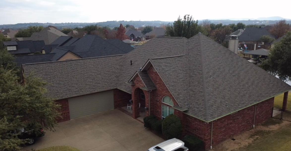 Aerial view of a brick house with a gray tiled roof, attached to a garage, surrounded by other houses and trees in a suburban neighborhood.