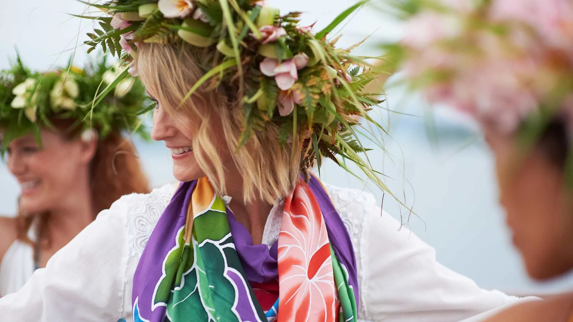 Women participating in a celebration or festival, wearing floral crowns and colorful scarves, smiling.
