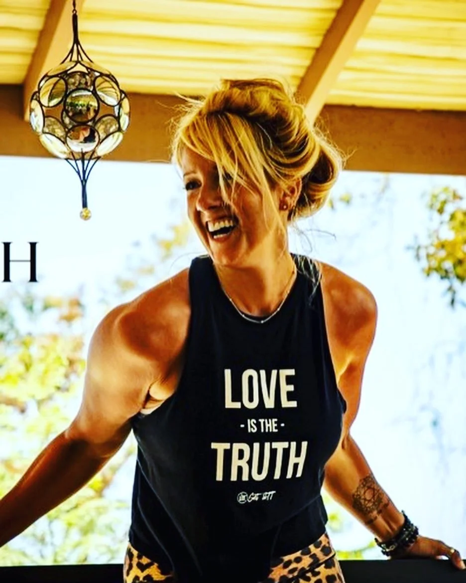 A woman with blonde hair styled in a messy bun, smiling and leaning forward, wearing a black sleeveless top with the text 'Love is the Truth' and leopard print pants, standing outdoors with trees in the background and a hanging decorative lantern overhead.