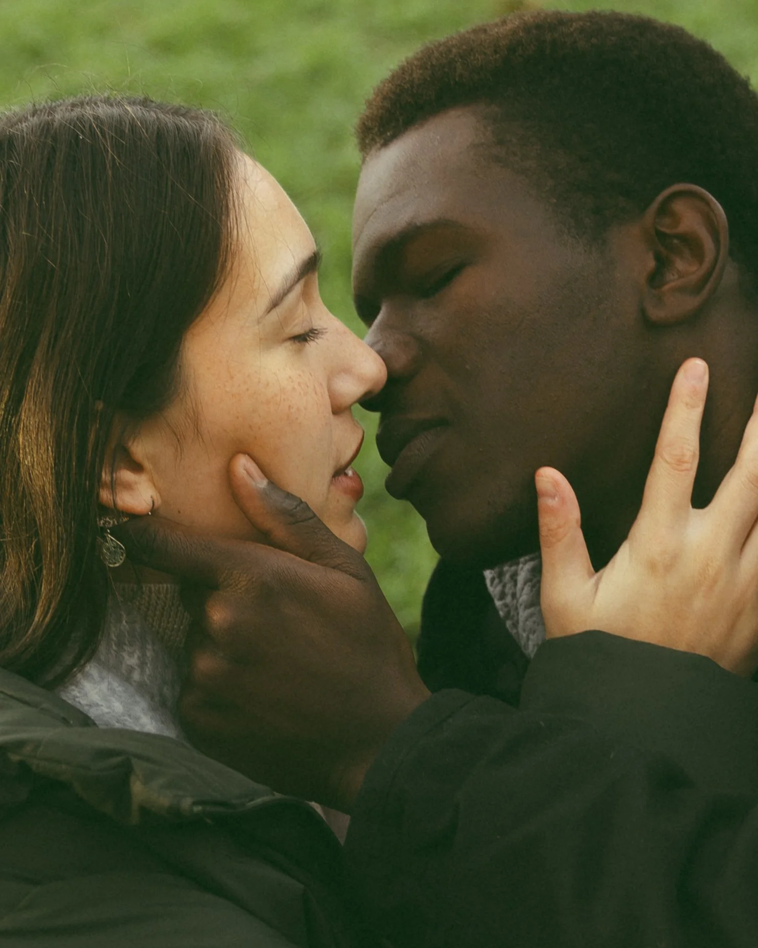 A close-up of a couple touching foreheads with eyes closed, outdoors with a grass background, showing intimacy and affection.