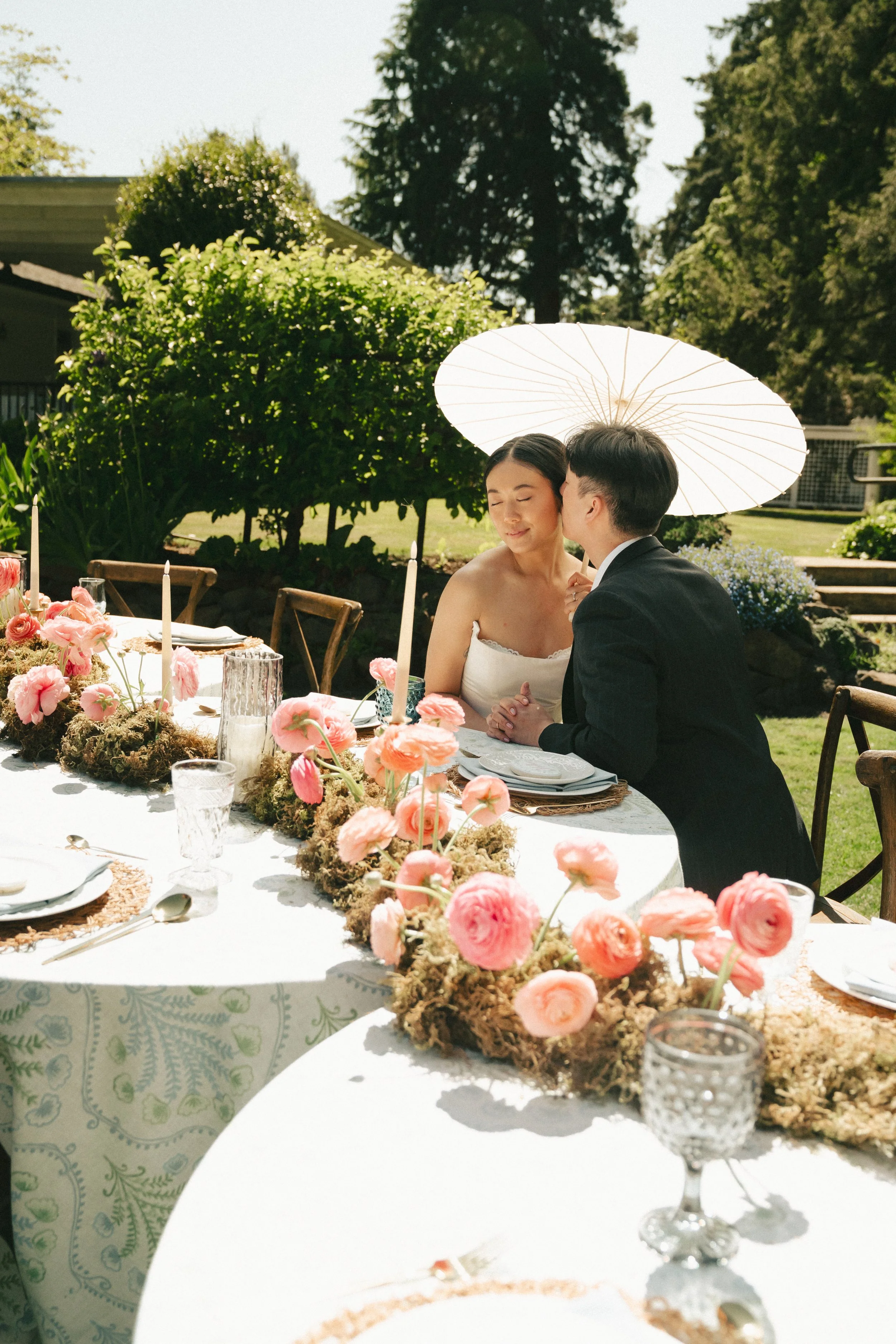 A man and woman sit closely at a wedding reception table outside under a large white umbrella, with the man kissing the woman on the cheek. The table is decorated with pink flowers, candles, and glassware.