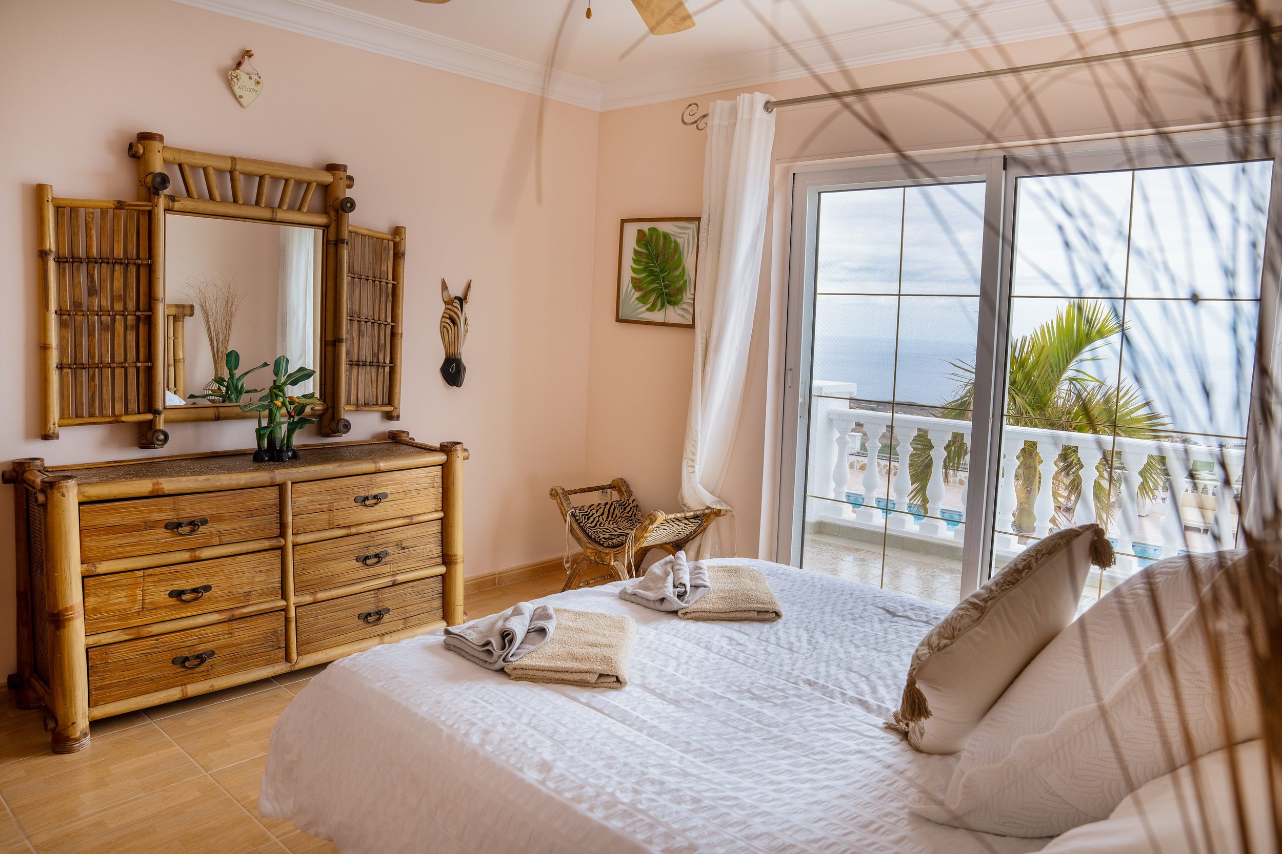 Bedroom with bamboo dresser, bed with white linens, towels, and slippers, framed leaf art, nearby balcony with view of palm trees and blue sky.