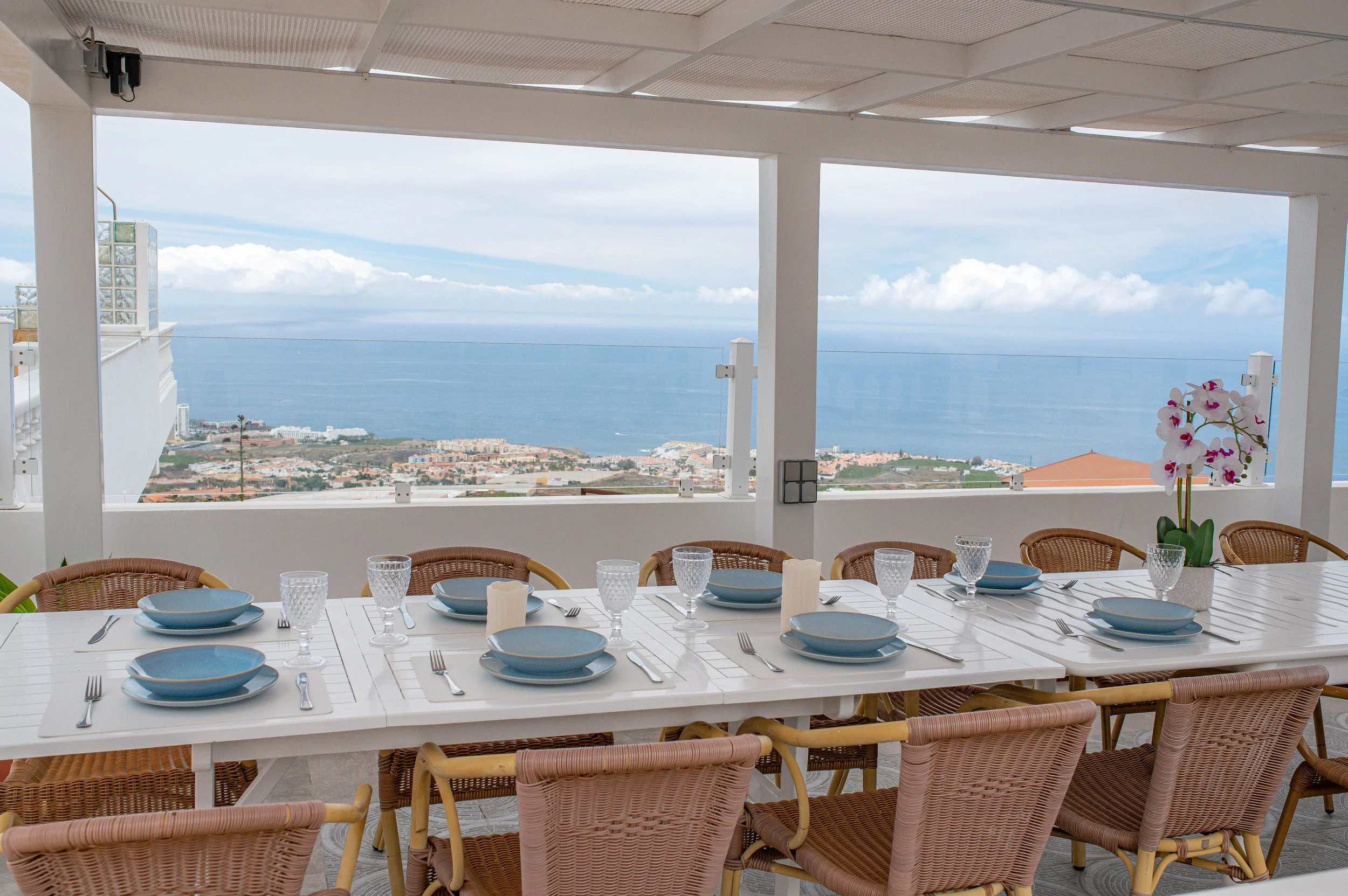 A dining table set with blue plates, glasses, and silverware, and a centerpiece with white orchids, overlooking a scenic view of the ocean and sky from a balcony or terrace with a white railing and columns.