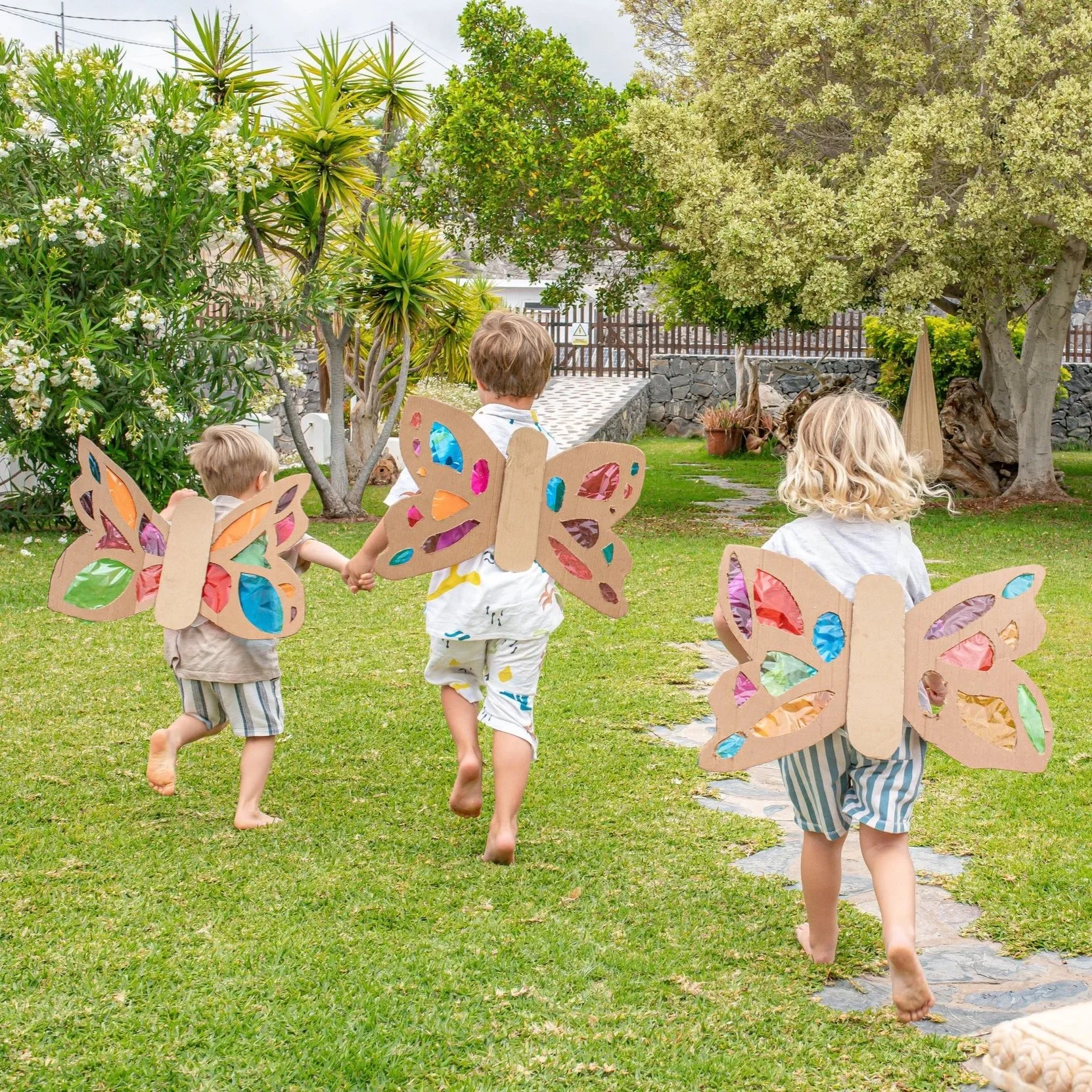 Three children in butterfly costumes running on grass in a backyard garden with trees, plants, and a stone wall.