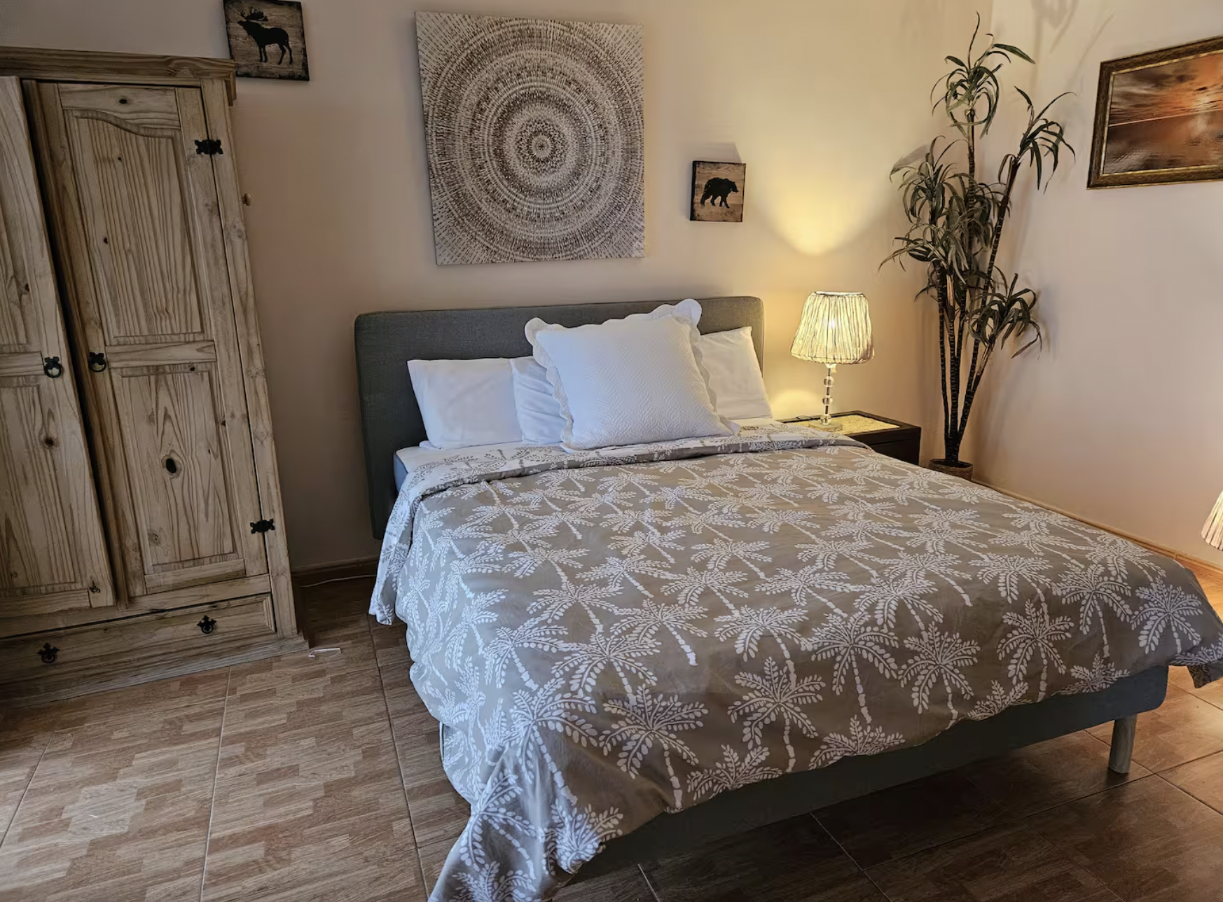 A bedroom with a bed, decorated with a patterned beige bedspread and white pillows. There is a grey headboard, a wooden wardrobe on the left, and a bedside table with a lamp on the right. The wall above the bed has artwork, including a large circular abstract piece, two smaller pictures of animals, and a sunset painting. A tall potted plant is next to the bedside table.