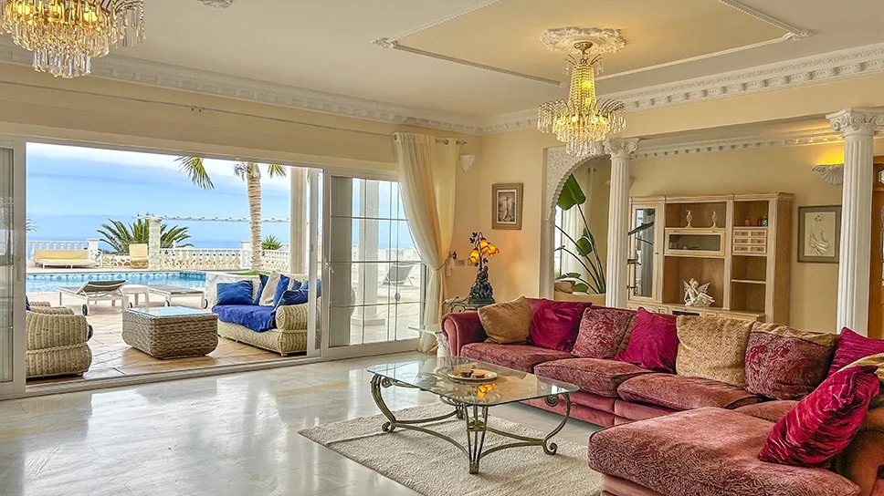 Living room with large sliding glass doors opening to a patio with seating, overlooking a swimming pool and the ocean, decorated with chandeliers and elegant furniture.