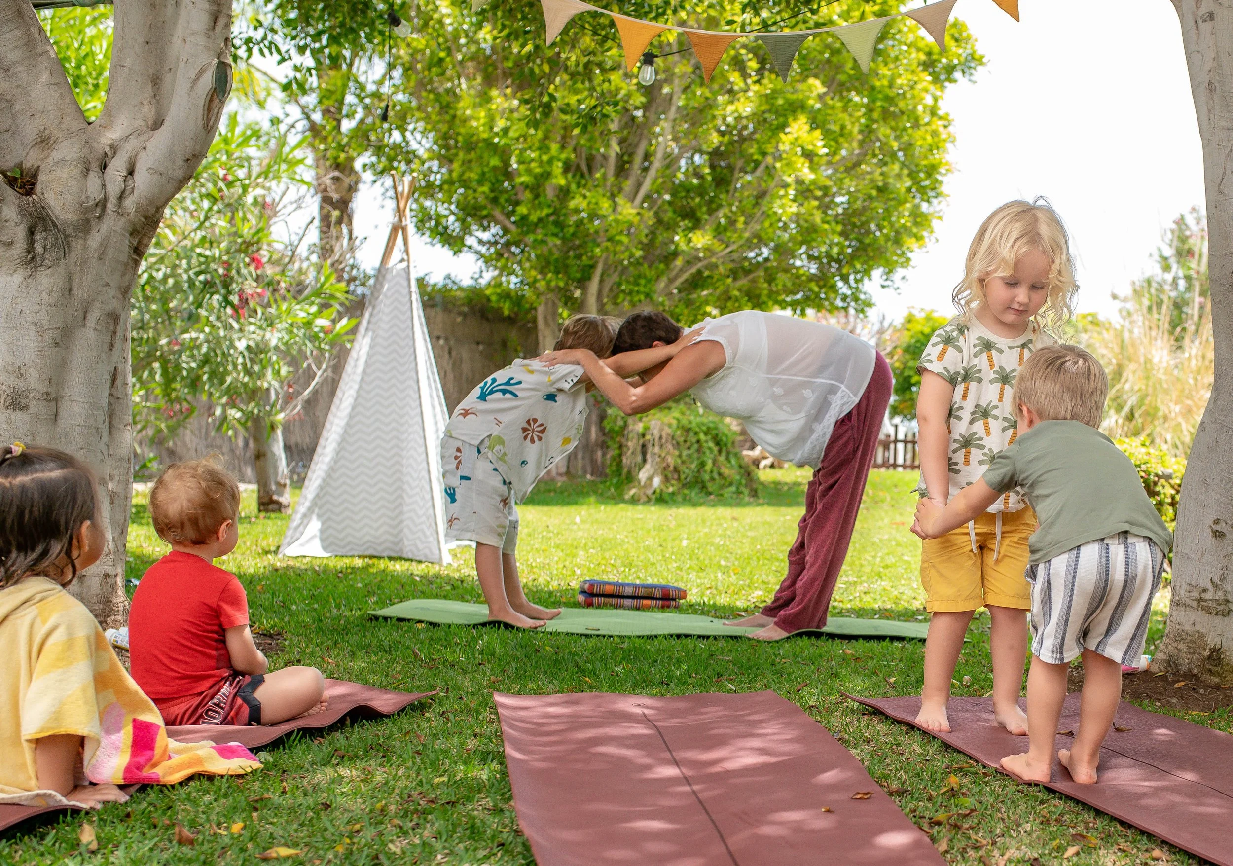 Children participating in a yoga or stretching class outdoors, with some sitting on mats and others standing in a tree pose, under shade trees with bunting decorations hanging overhead.