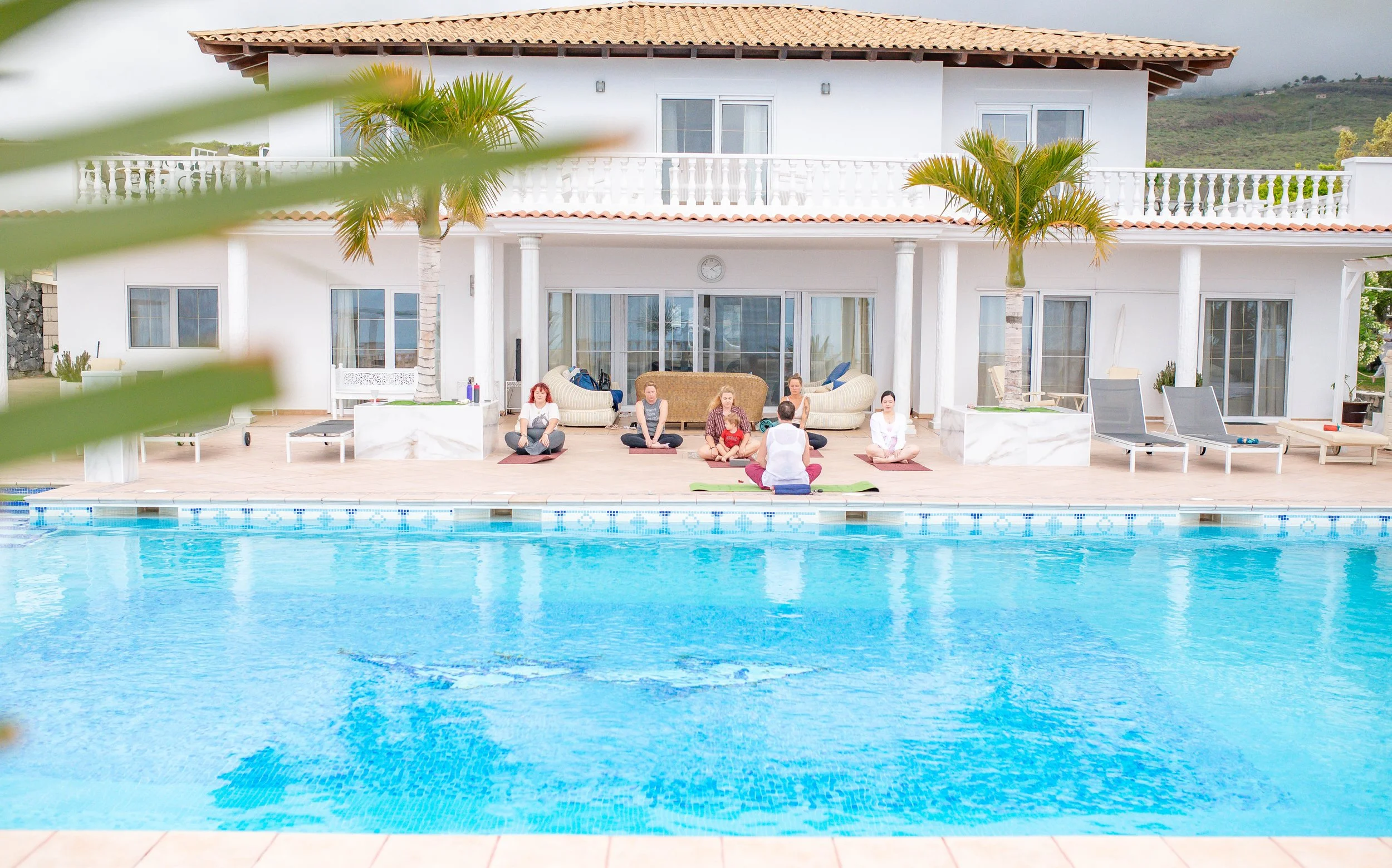 People practicing yoga by a swimming pool at a resort with palm trees and a white building in the background.
