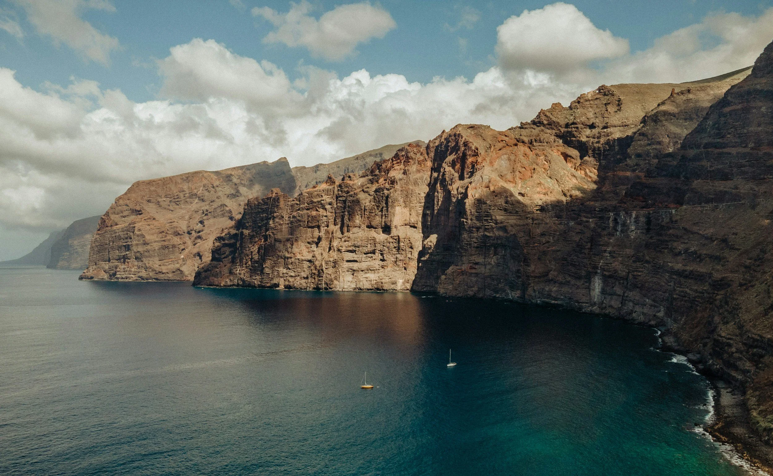 Cliffs along a coastline with water below, a partly cloudy sky, and two sailboats in the water.