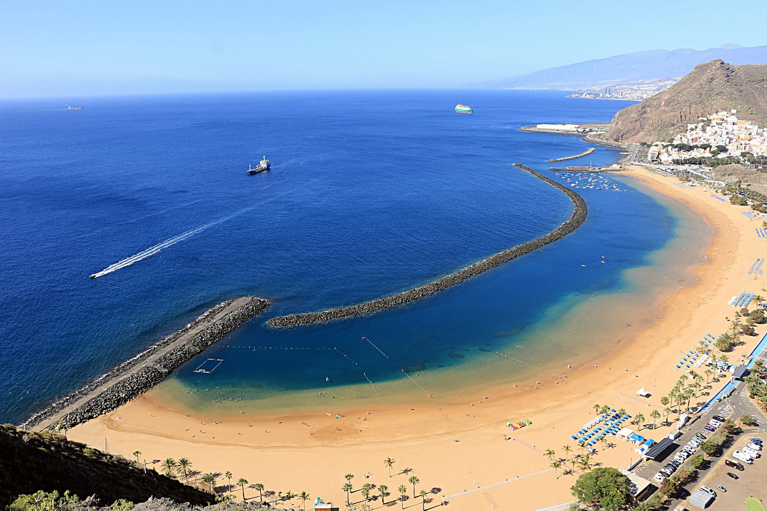 Aerial view of a beach with golden sand and turquoise water bordered by a row of palm trees and colorful umbrellas, with boats and ships in the distant ocean and a hillside with a town and mountains in the background.