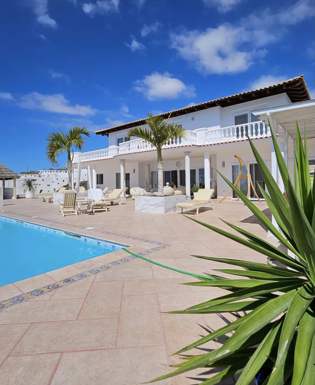A luxurious outdoor pool area with lounge chairs, palm trees, and a white two-story house under a partly cloudy blue sky.
