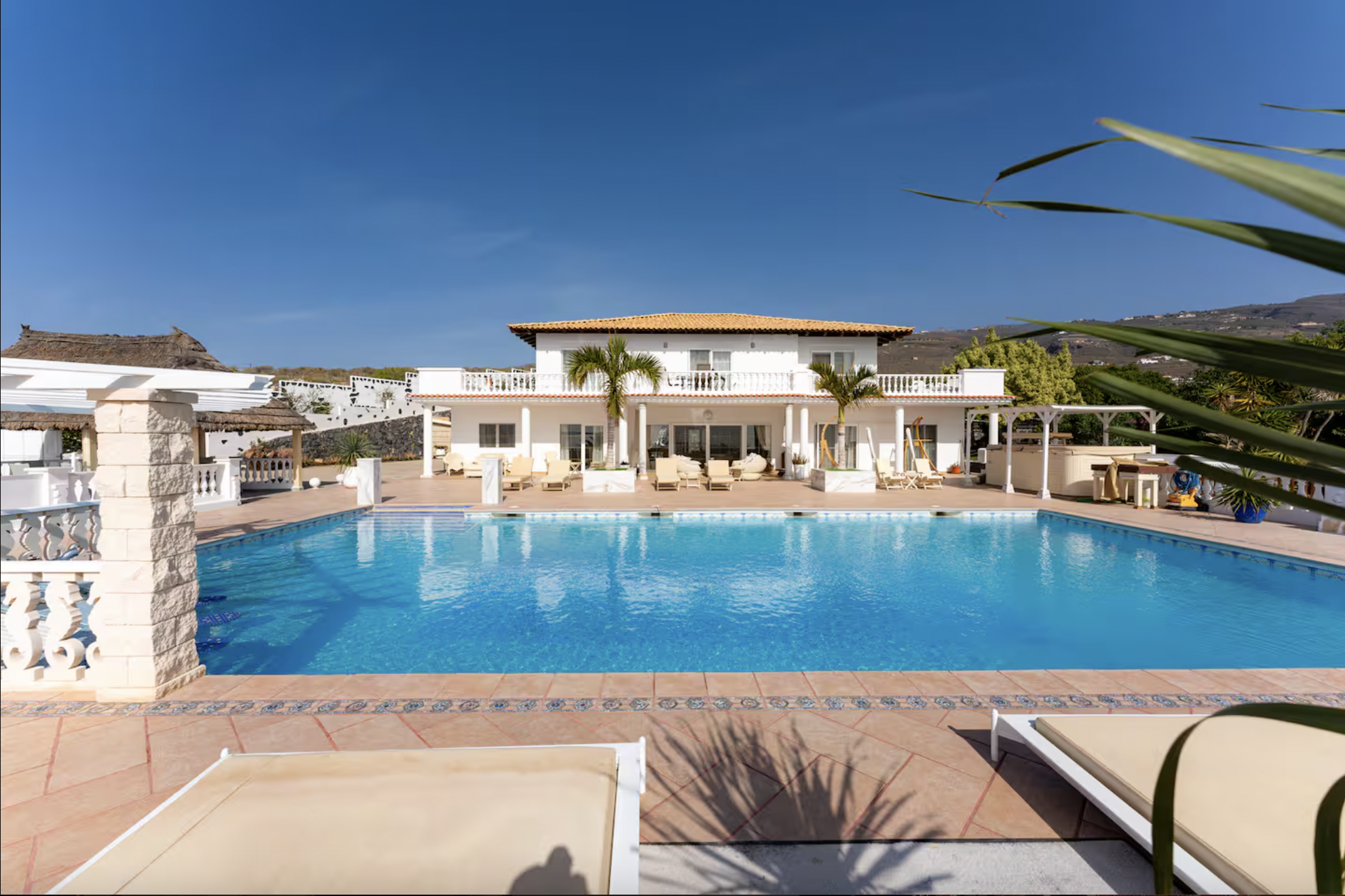 Luxury villa with a swimming pool, outdoor lounge chairs, palm trees, and a white building with balconies under a blue sky.