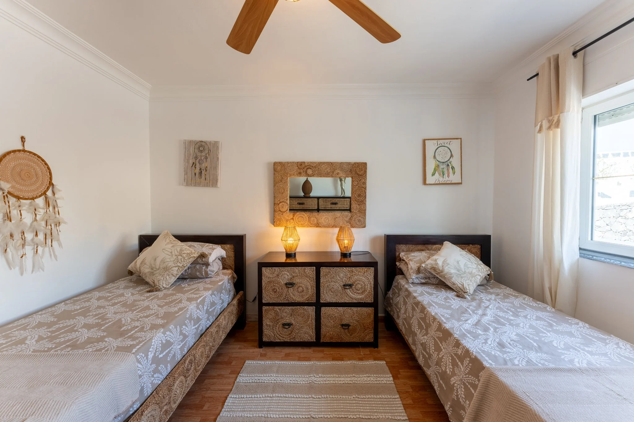 A bedroom with two single beds, each with patterned bedspreads and pillows, a wooden dresser with woven drawers and two lamps, decorated with wall art, a mirror, and a window with curtains.