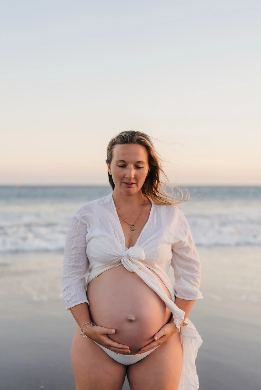 A pregnant woman standing on the beach with ocean waves in the background, wearing a white tied shirt and white underwear, holding her belly and looking down.