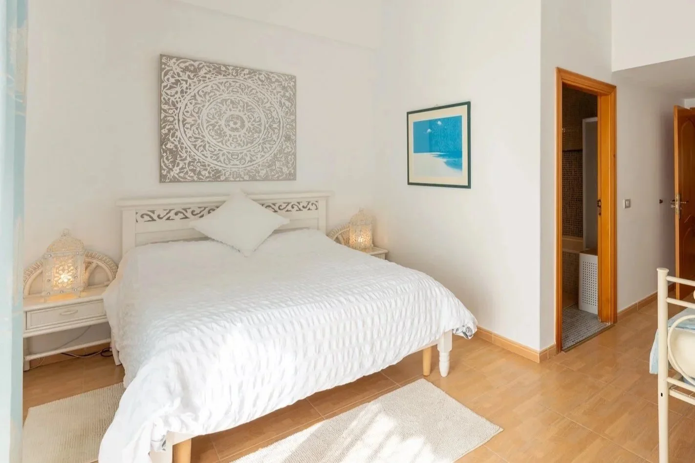 A cozy bedroom with a white bed, decorative pillows, wall art, nightstands with lamps, and an open doorway showing a bathroom with tiled walls.