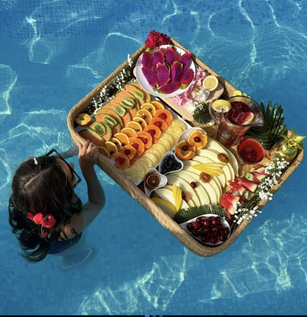 A young girl with dark hair and a pink flower hair clip reaches for a floating tray of assorted fresh fruits and drinks in a swimming pool.