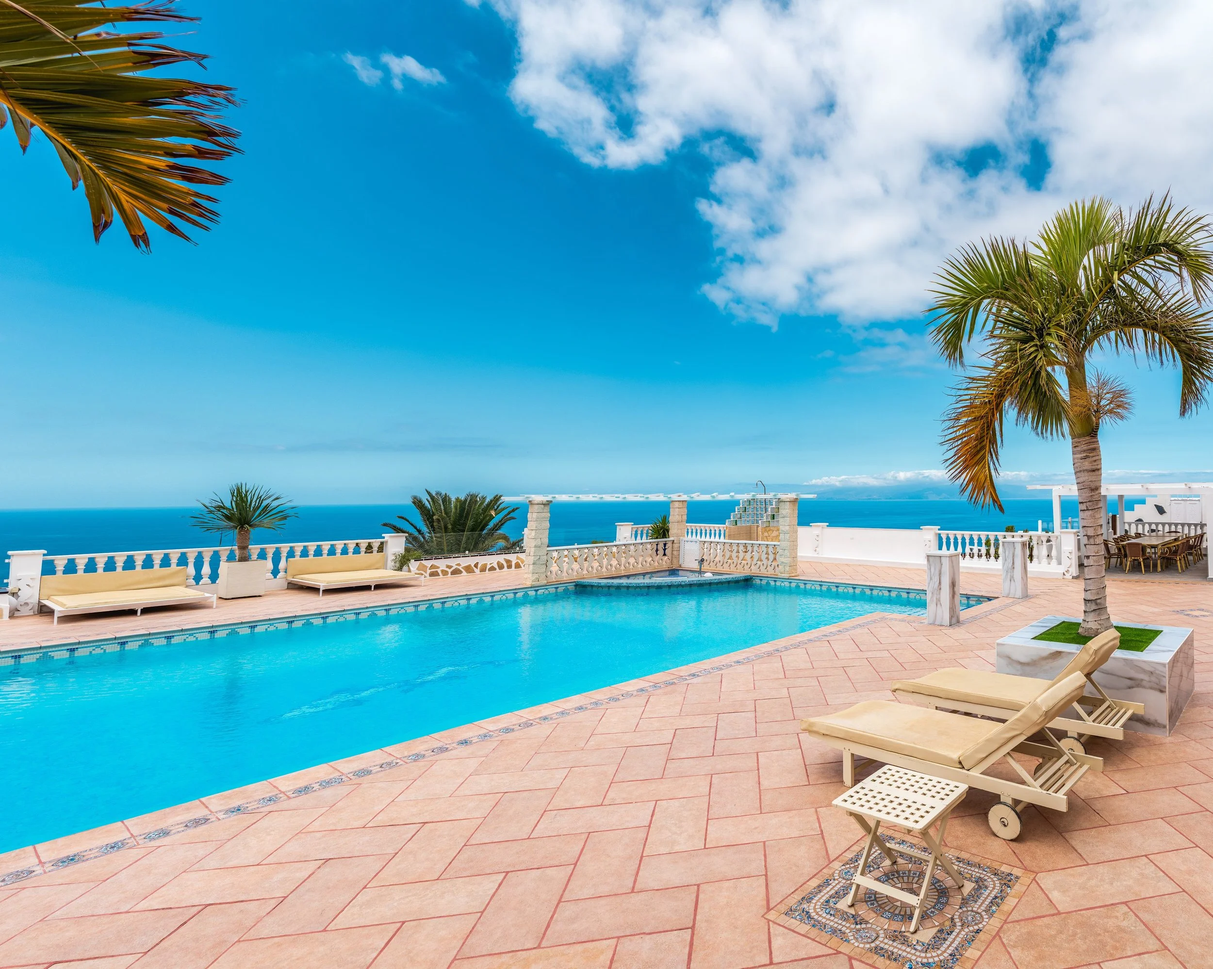 A view of a swimming pool area with beige lounge chairs, palm trees, and a background of ocean and blue sky with clouds.