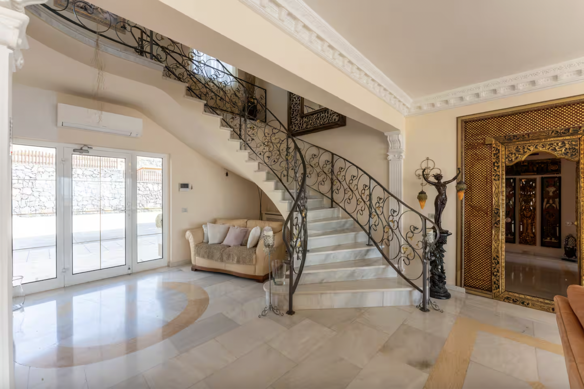 Living room with large windows, glass doors, a staircase with ornate iron railing, a beige sofa with throw pillows, a decorative statue, and intricate crown molding.