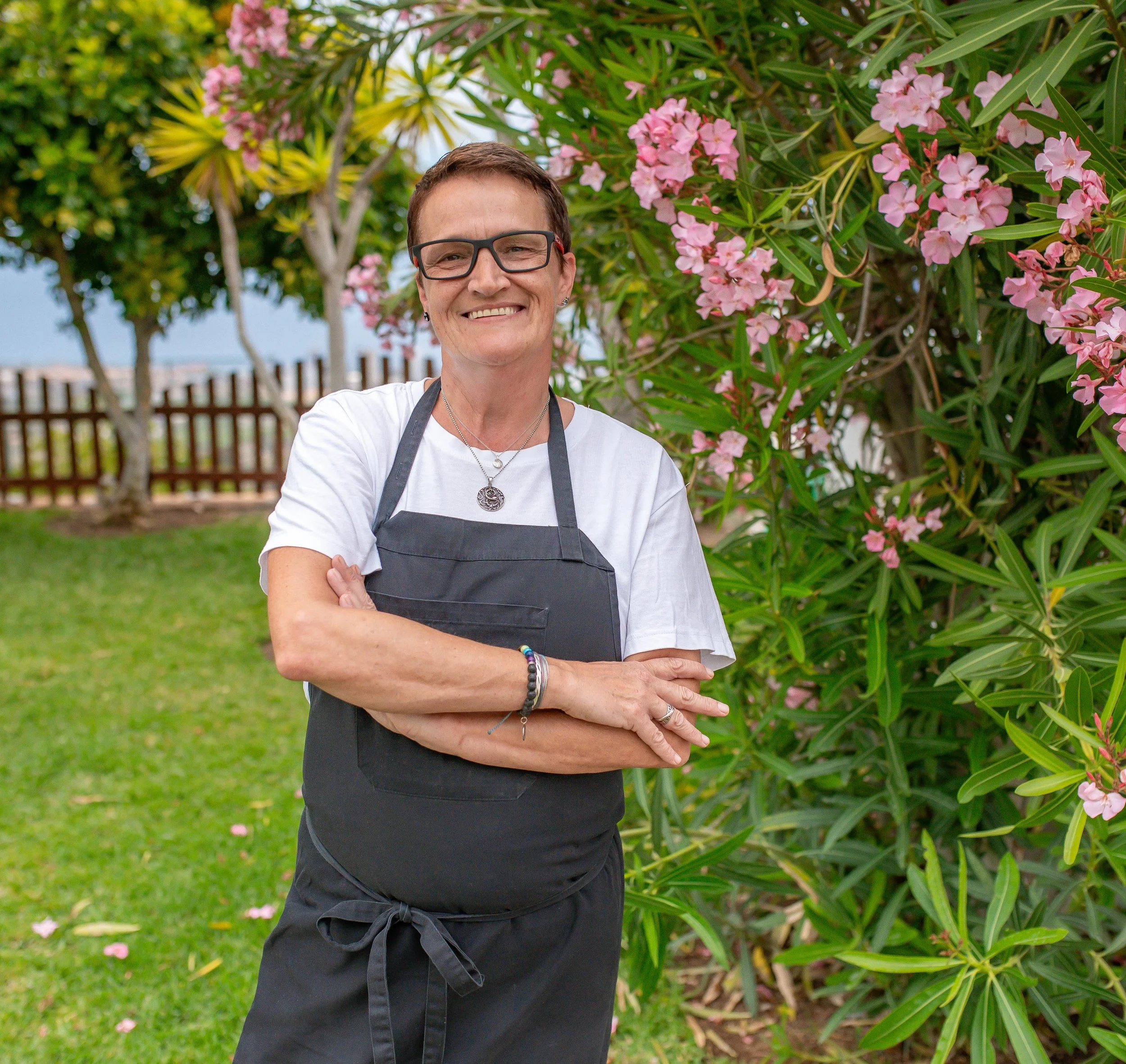 A smiling person with short brown hair, wearing glasses, a white t-shirt, a black apron, and jewelry, standing outdoors in front of flowering bushes with pink flowers and green leaves.
