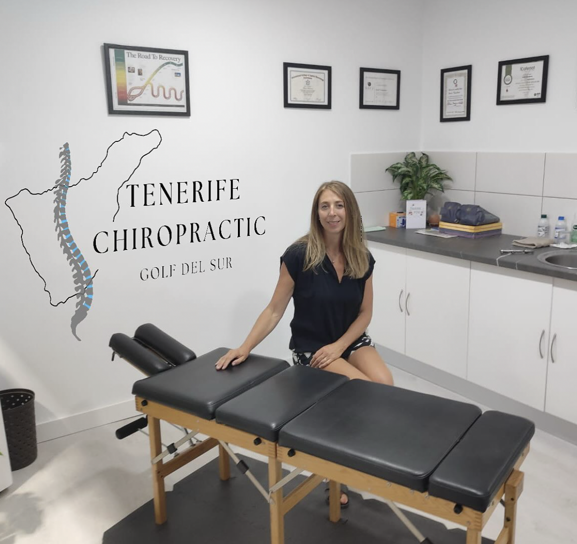 Woman sitting on a chiropractic massage table in an office with framed certificates on the wall and a sign saying 'Tenerife Chiropractic Golf del Sur' with a map of Tenerife.