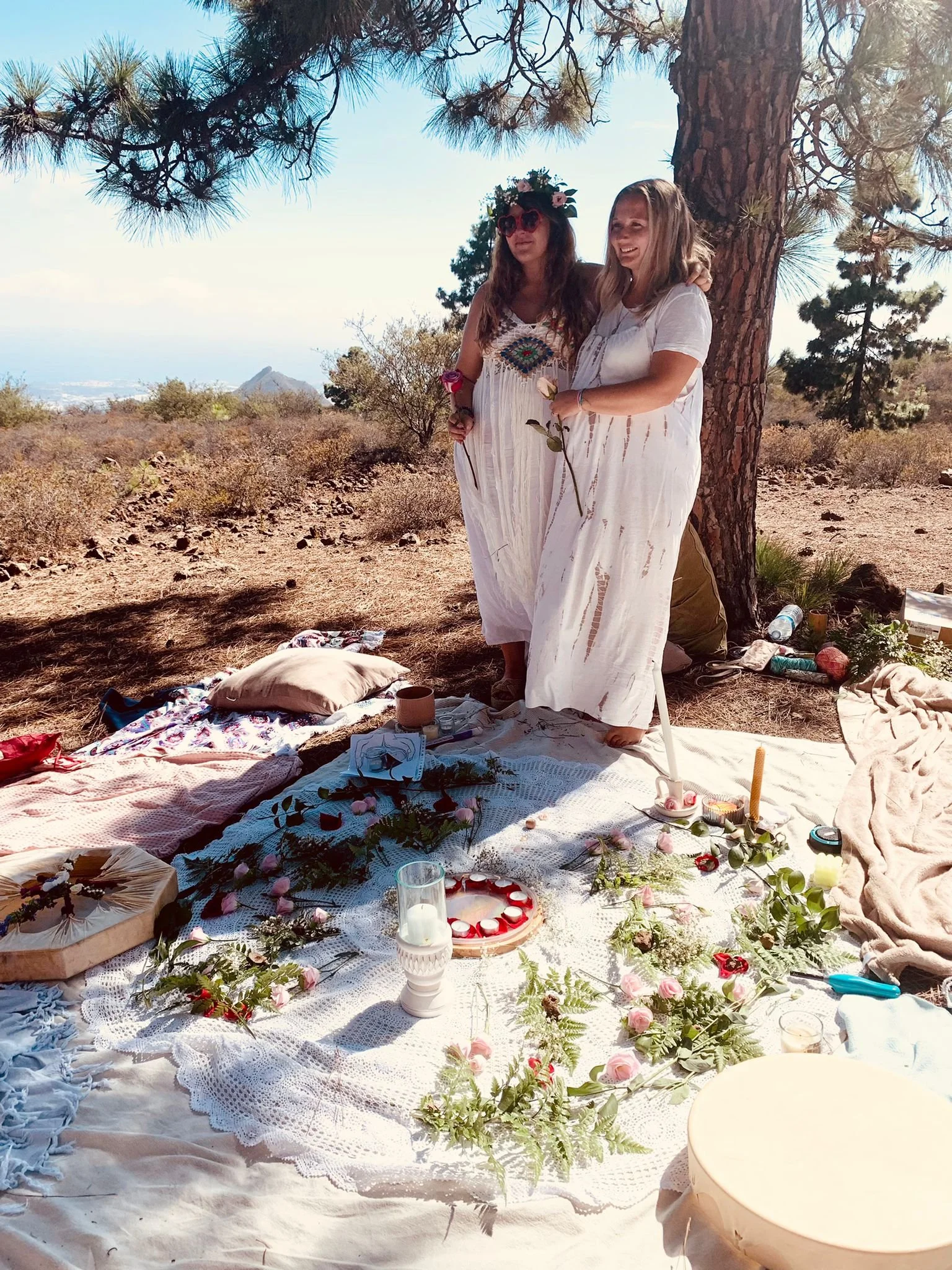 Two women dressed in white standing under a large tree during a daytime outdoor gathering, with a decorated blanket and various items like candles, flowers, and cups on the blanket.