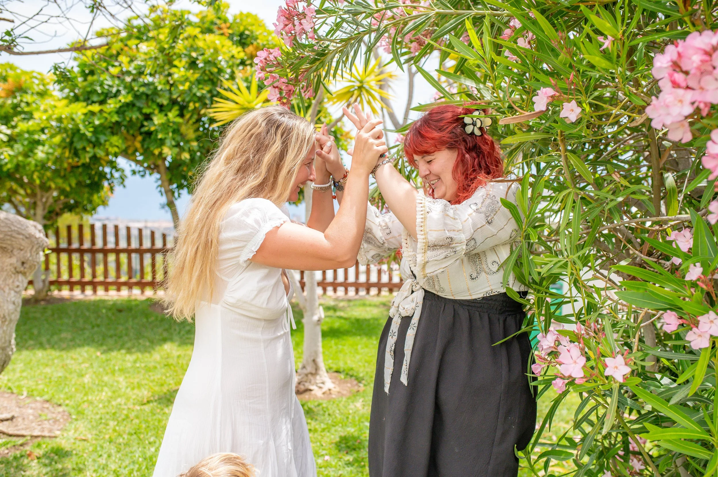 Two women with long hair smiling and playfully holding hands together in a garden, surrounded by green trees and pink flowers.