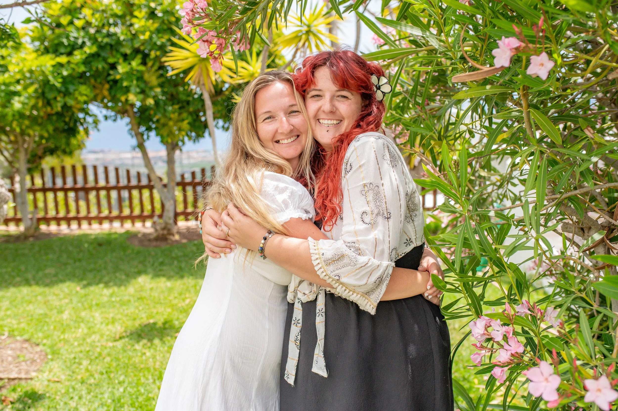 Two women hugging and smiling outdoors amidst greenery and pink flowers, with a wooden fence and city in the background.