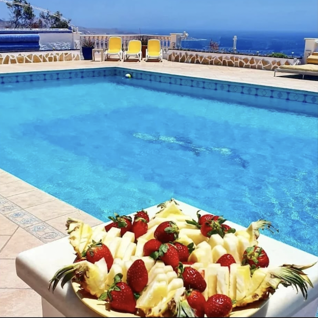 A swimming pool on a sunny day with patio chairs and a scenic ocean view in the background; a plate of sliced pineapple and strawberries in the foreground.