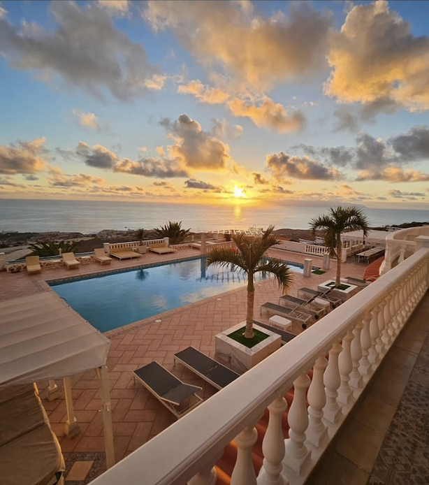 View of a swimming pool on a terrace overlooking the ocean during sunset, with lounge chairs, palm trees, and a decorative railing.