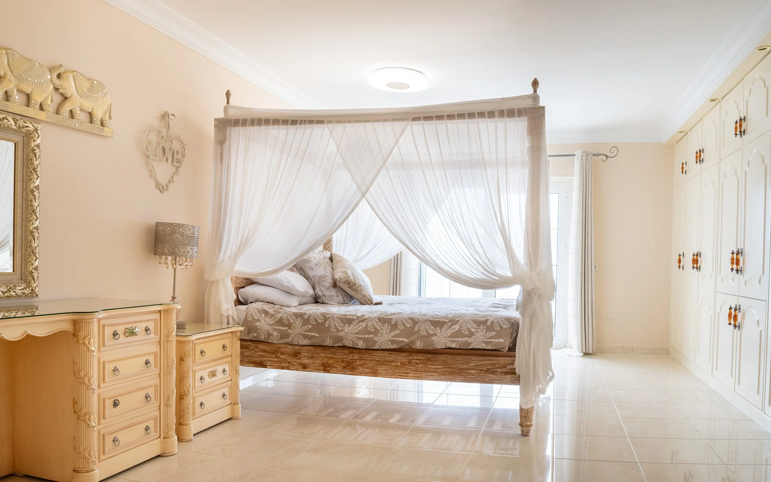 A bedroom with a four-poster bed with sheer white curtains, beige and white bedding, a beige nightstand with decorative drawers, a lamp, a mirror, and a wall hanging with the word "LOVE" inside a heart shape.