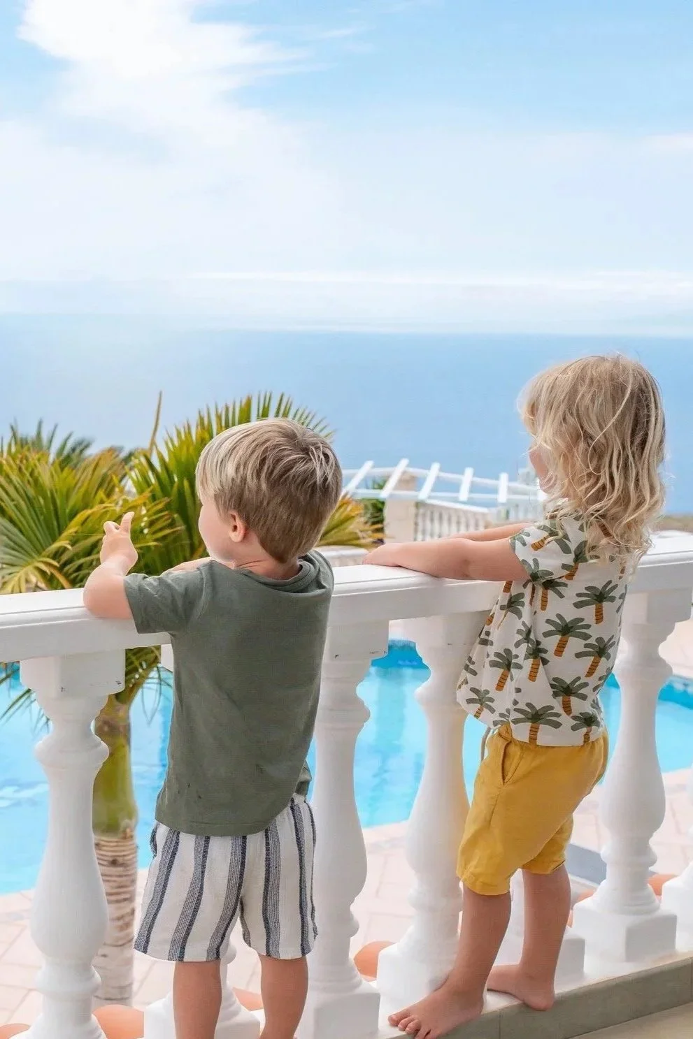 Two young children, a boy and a girl, standing on a balcony overlooking the ocean with lush greenery and a partly cloudy sky in the background.