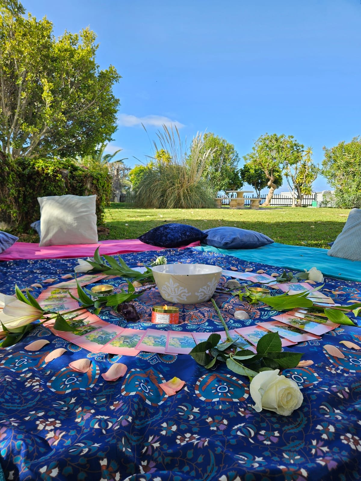 A circular arrangement of tarot cards, a white bowl, flowers, and candles on a colorful cloth outdoors on a grassy area with trees and a clear blue sky in the background.