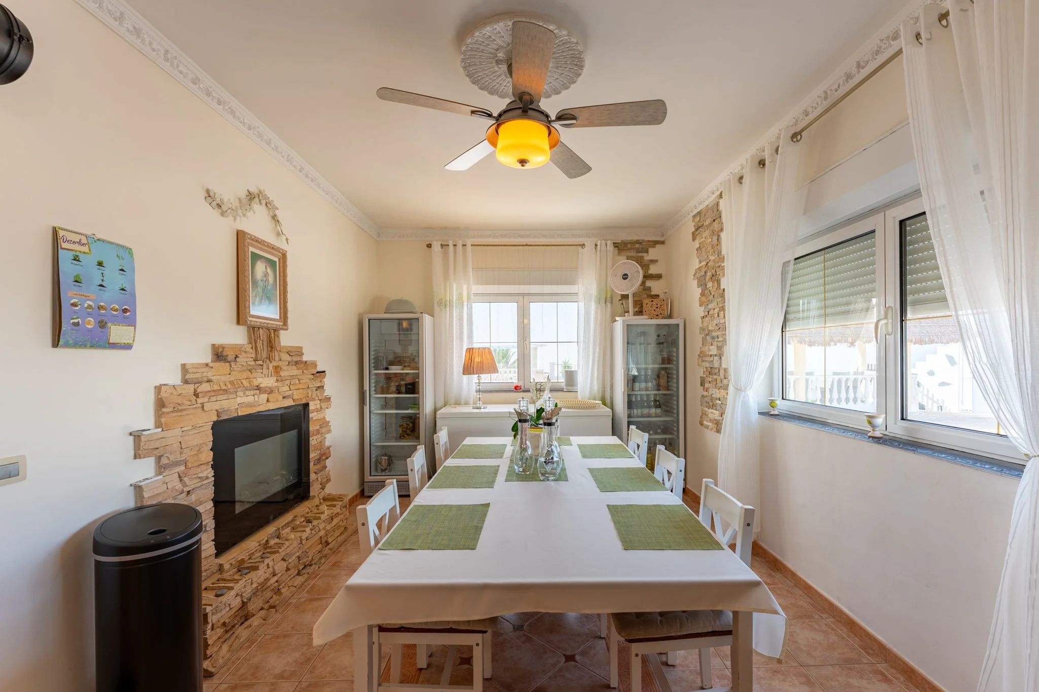 Dining room with a table set with a white tablecloth and green placemats, surrounded by white chairs. The room has large windows with white curtains, a ceiling fan with lights, a stone accent wall, and a corner display cabinet.