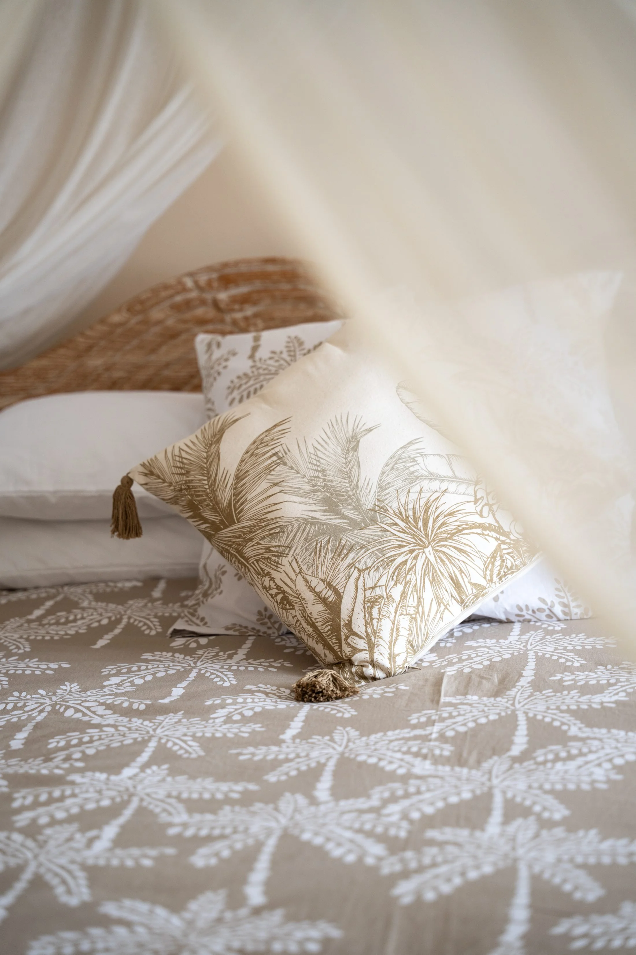 A beige tablecloth with a white leafy pattern, a decorative pillow with gold palm tree prints and tassels, and a corner of an open book with white pages.