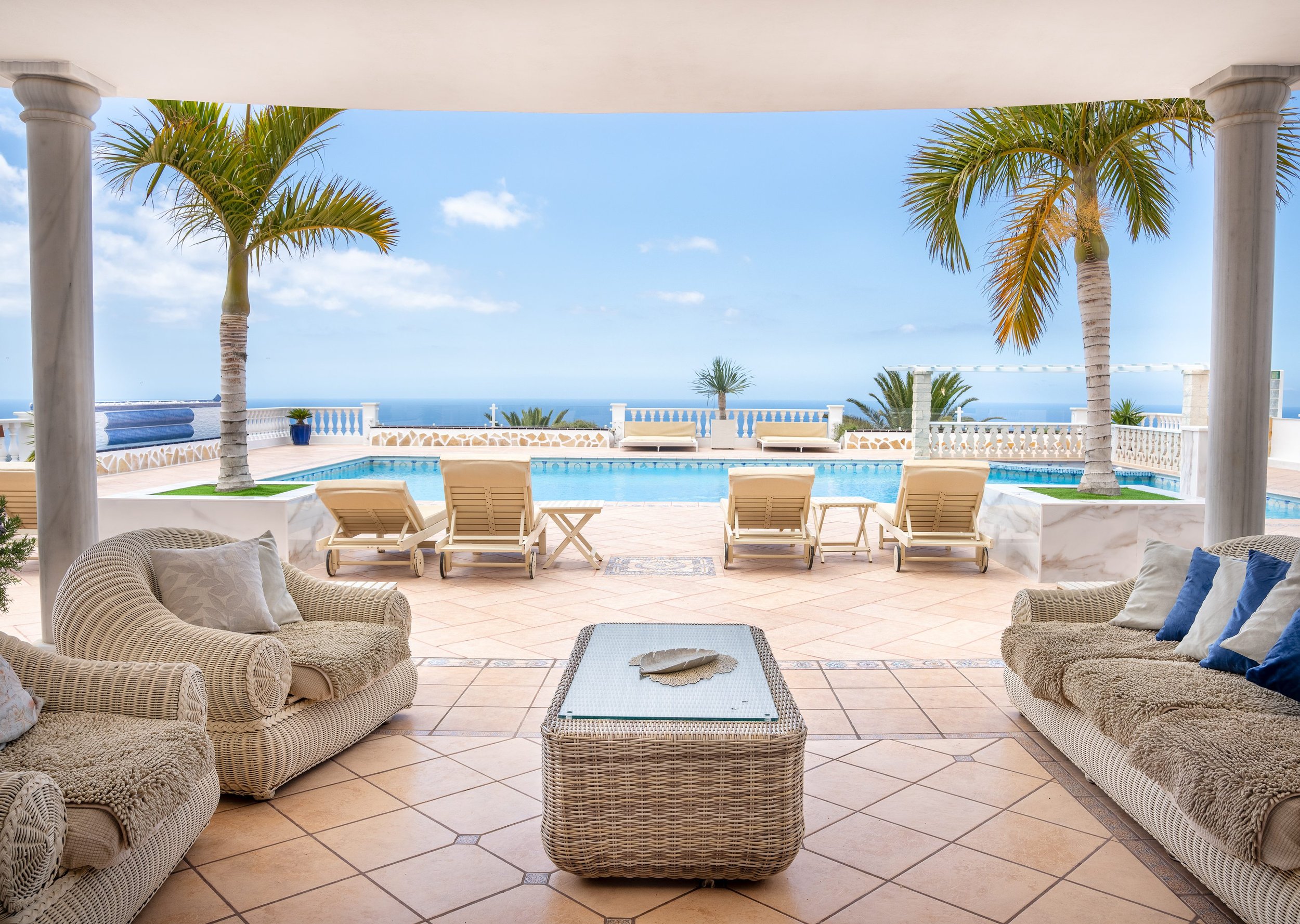 View of a poolside lounge area with wicker chairs, a coffee table, and palm trees overlooking the ocean, seen from a covered patio with columns.