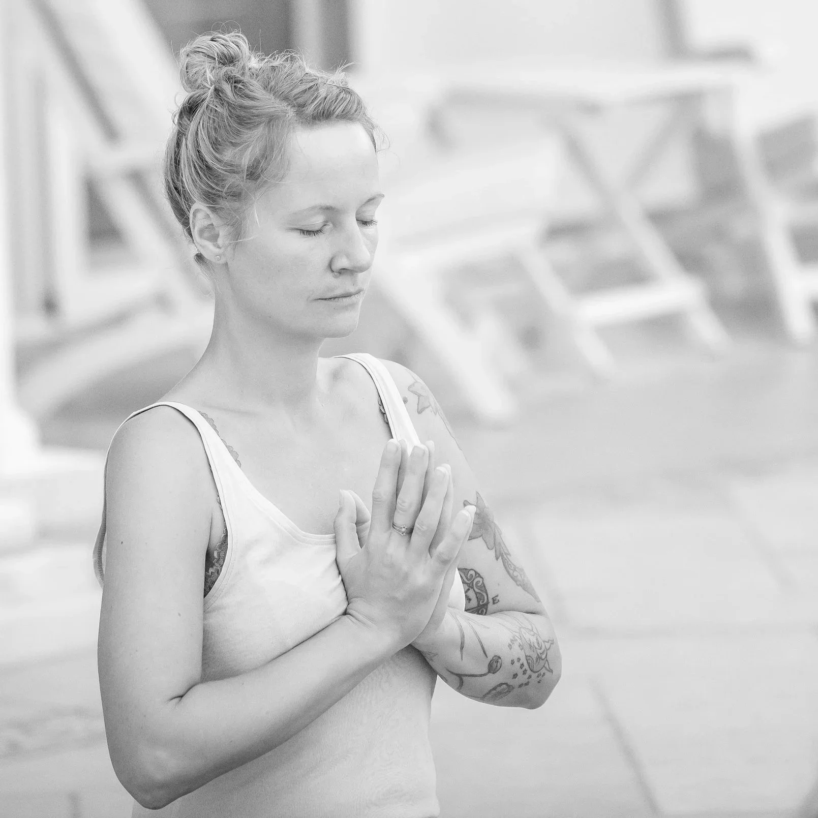 A woman with tattoos practicing yoga outdoors, with eyes closed and hands pressed together in a prayer position.