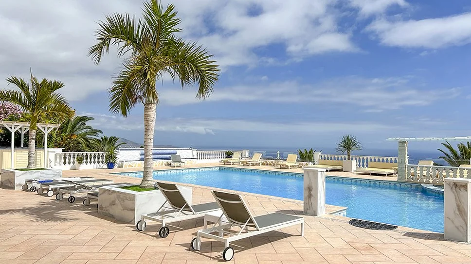 A luxurious pool area with palm trees, lounge chairs, and a view of the ocean under a partly cloudy sky.
