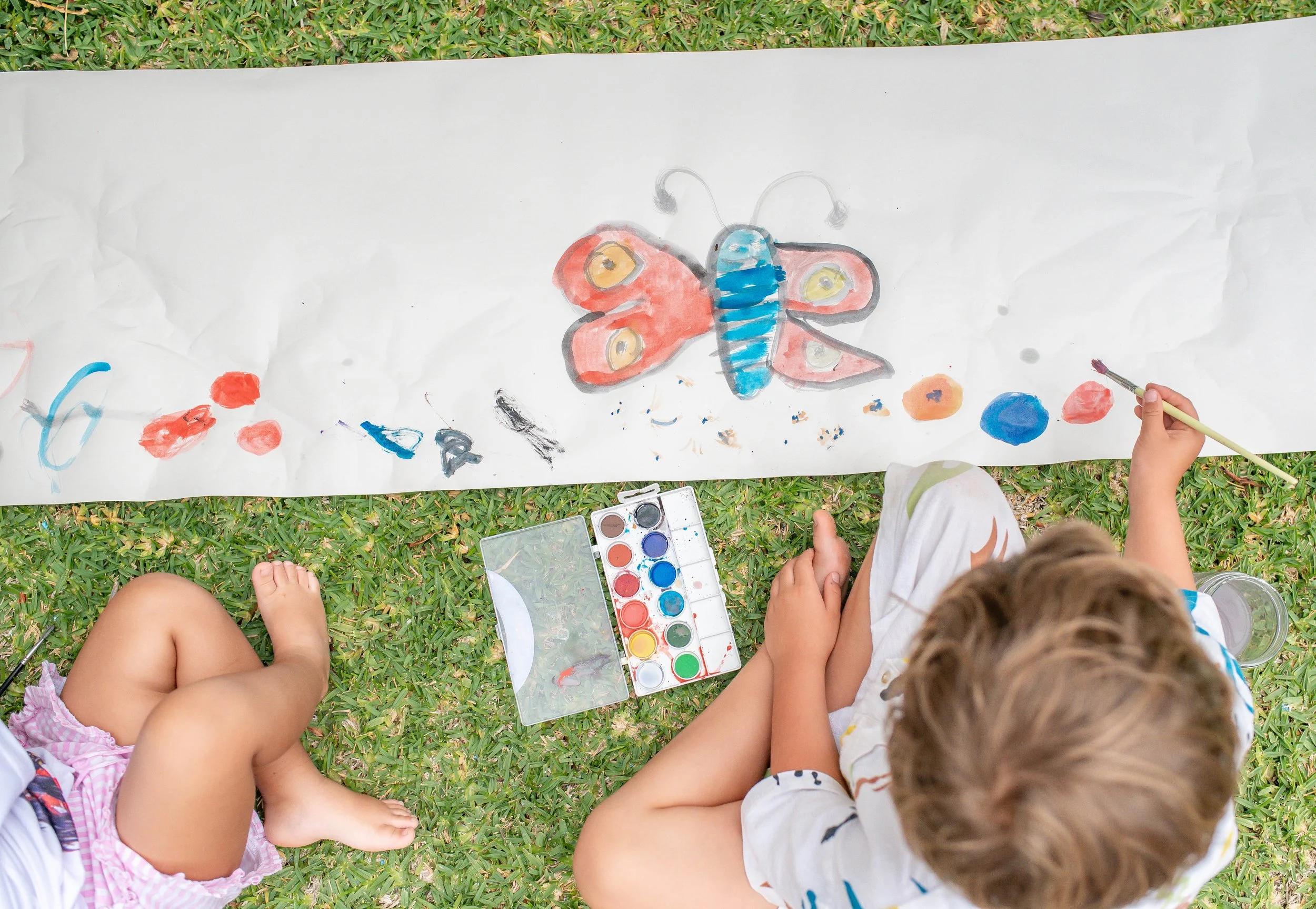 Children sitting on grass around a long sheet of paper with a colorful butterfly painting, watercolor paints, and a paintbrush.