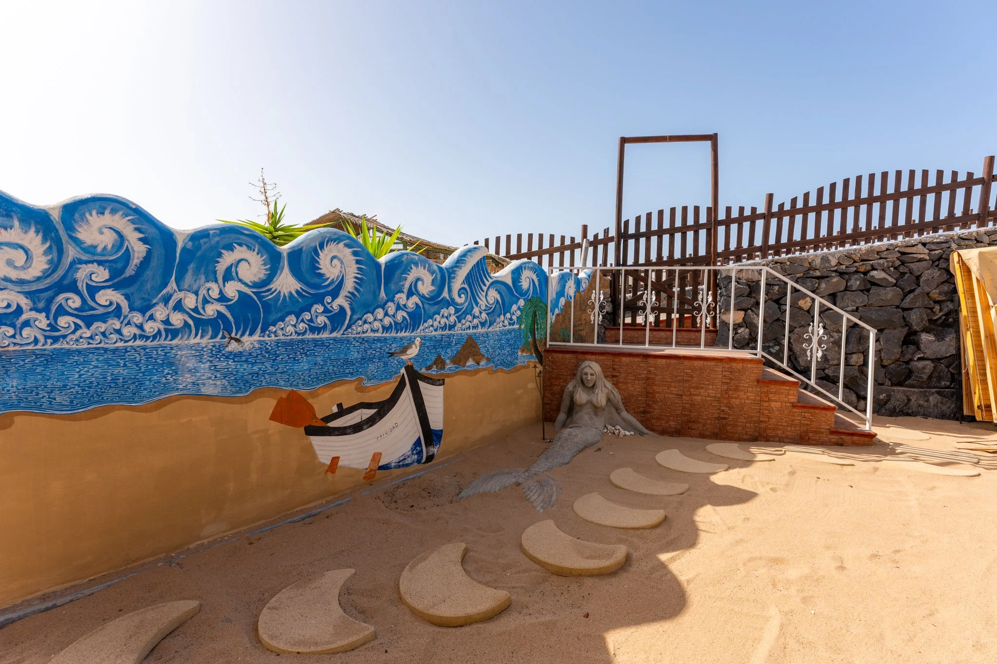 Colorful mural of ocean waves with dolphins, a boat, and greenery, adjacent to a set of decorative steps, with a statue of a mermaid at the base, and a stone fence in the background.