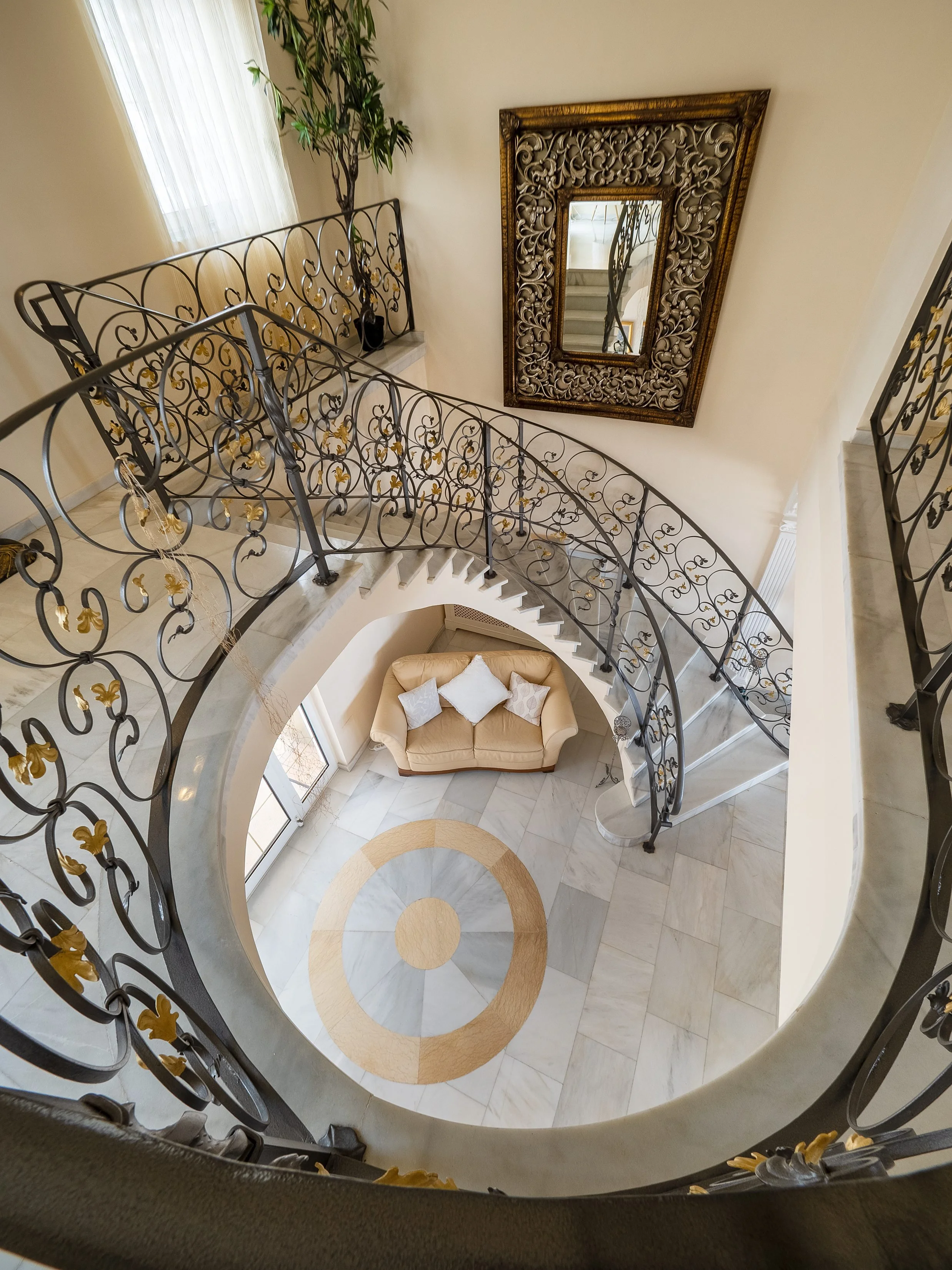 Looking down at a spiral staircase with ornate black wrought-iron railings decorated with gold accents. At the bottom, there's a small seating area with a beige curved sofa and three white pillows. The floor has a circular pattern with beige and white marble tiles, and the staircase leads to a second floor. There is a large mirror with an intricate gold frame on the wall, and a tall green potted plant near a window with white sheer curtains.