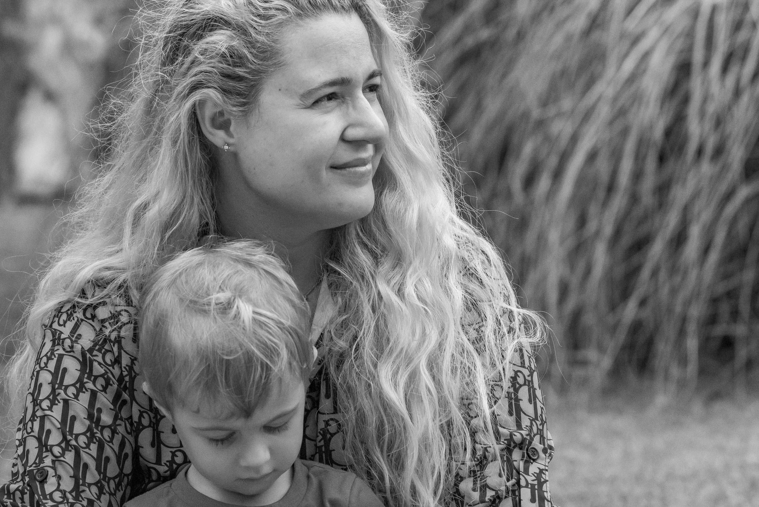 A woman with long, wavy blonde hair is looking to her left and smiling gently. She is wearing earrings and a patterned shirt. A young boy with short hair, closed eyes, and a neutral expression is resting his head on her chest or shoulder. The background is blurred with some tall grass or plants.