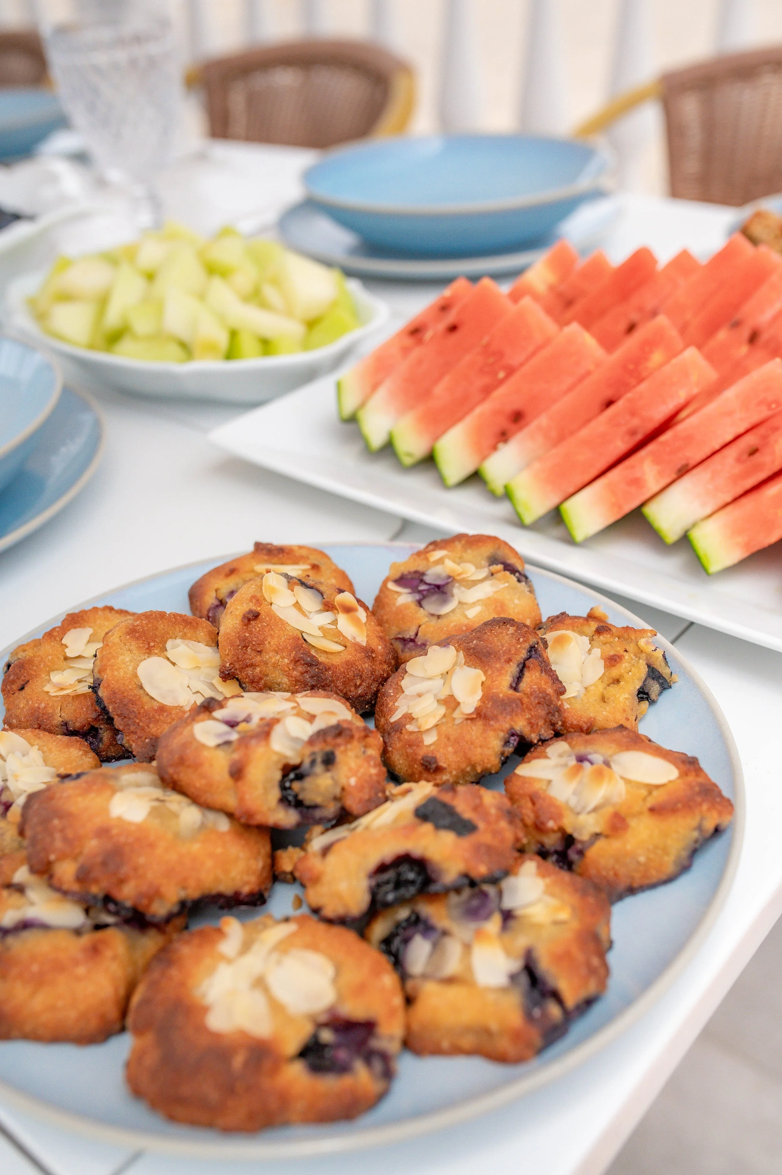 A plate of chocolate chip cookies topped with sliced almonds in the foreground. In the background, there is a white bowl filled with chopped honeydew melon and a blue plate with slices of watermelon.