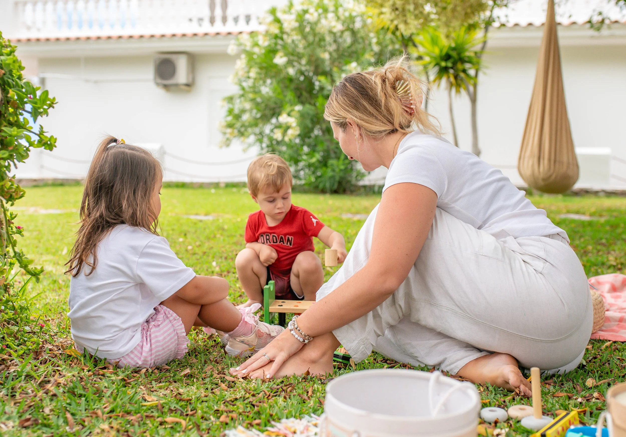 A woman and two young children sitting on the grass outdoors, engaging in a playful activity. The woman is wearing a white top and has blonde hair, while the children are dressed casually. There are trees, a building, and a large decorative hanging basket in the background.