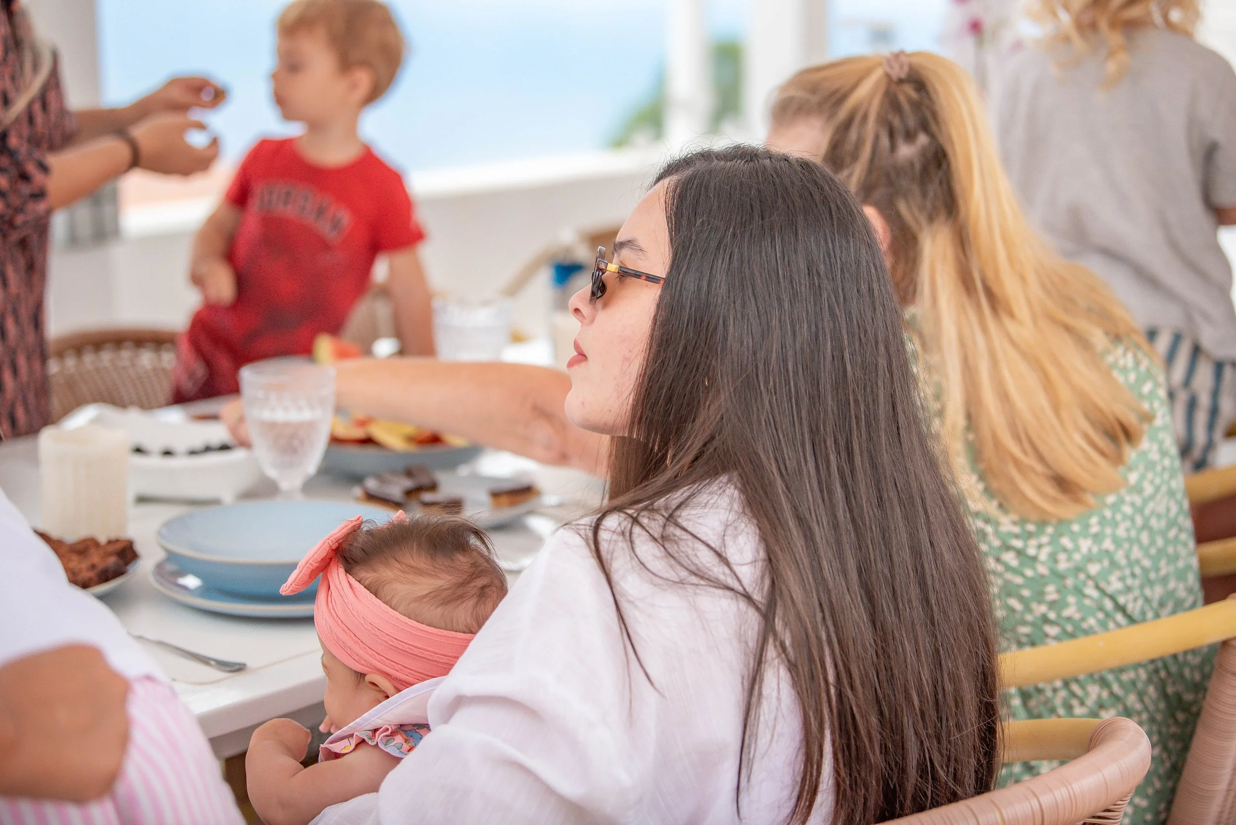 A woman with long dark hair and glasses holding a baby, seated at a table with food and drinks. Other people, including children, are present in the background, at what appears to be a celebration or gathering.