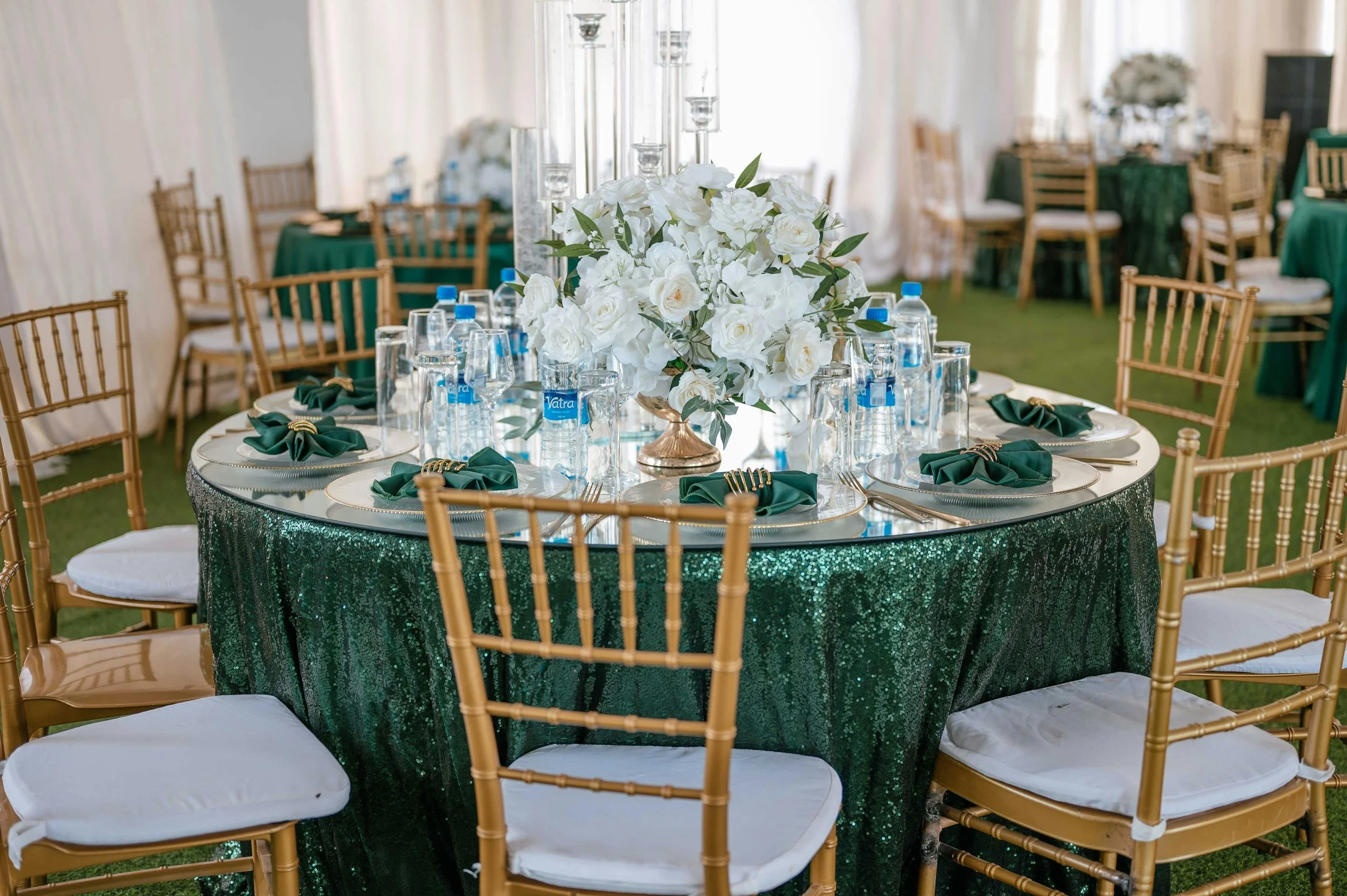 Wedding reception table with a large white floral centerpiece, surrounded by gold chairs with white cushions, green sequin tablecloth, set with glassware, green napkins, and gold flatware.