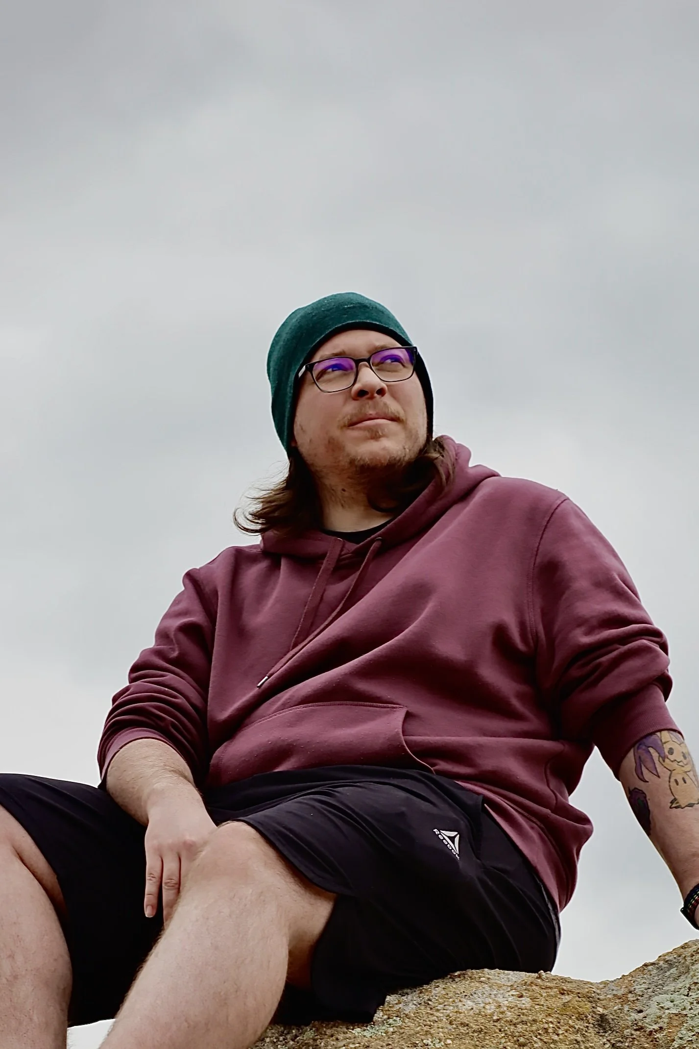 A man with glasses and a green beanie hat sitting on a rock under a cloudy sky, wearing a maroon hoodie and black shorts.